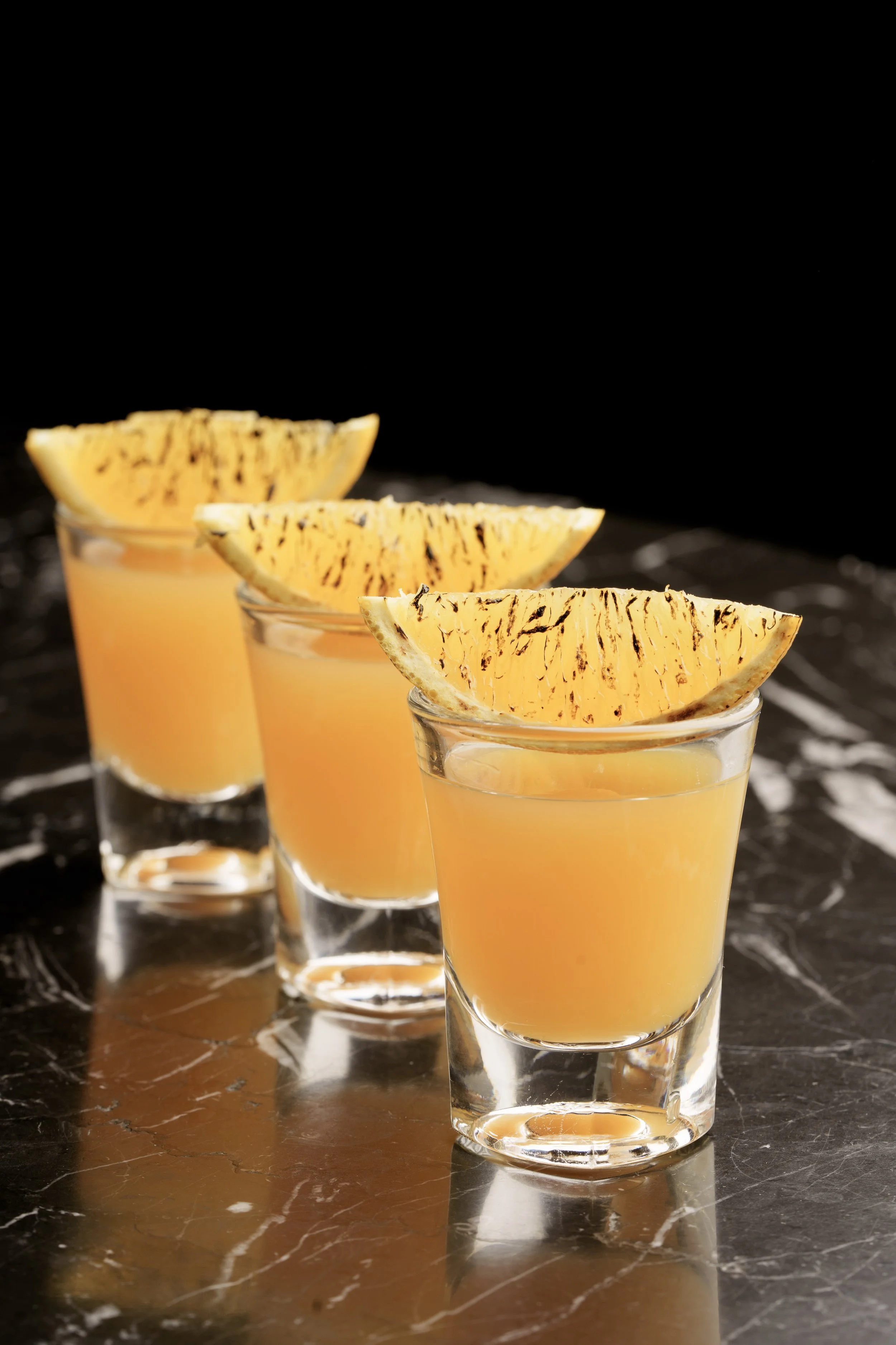 Three shot glasses filled with orange-colored beverage, garnished with charred lemon wedges on a dark marble surface.