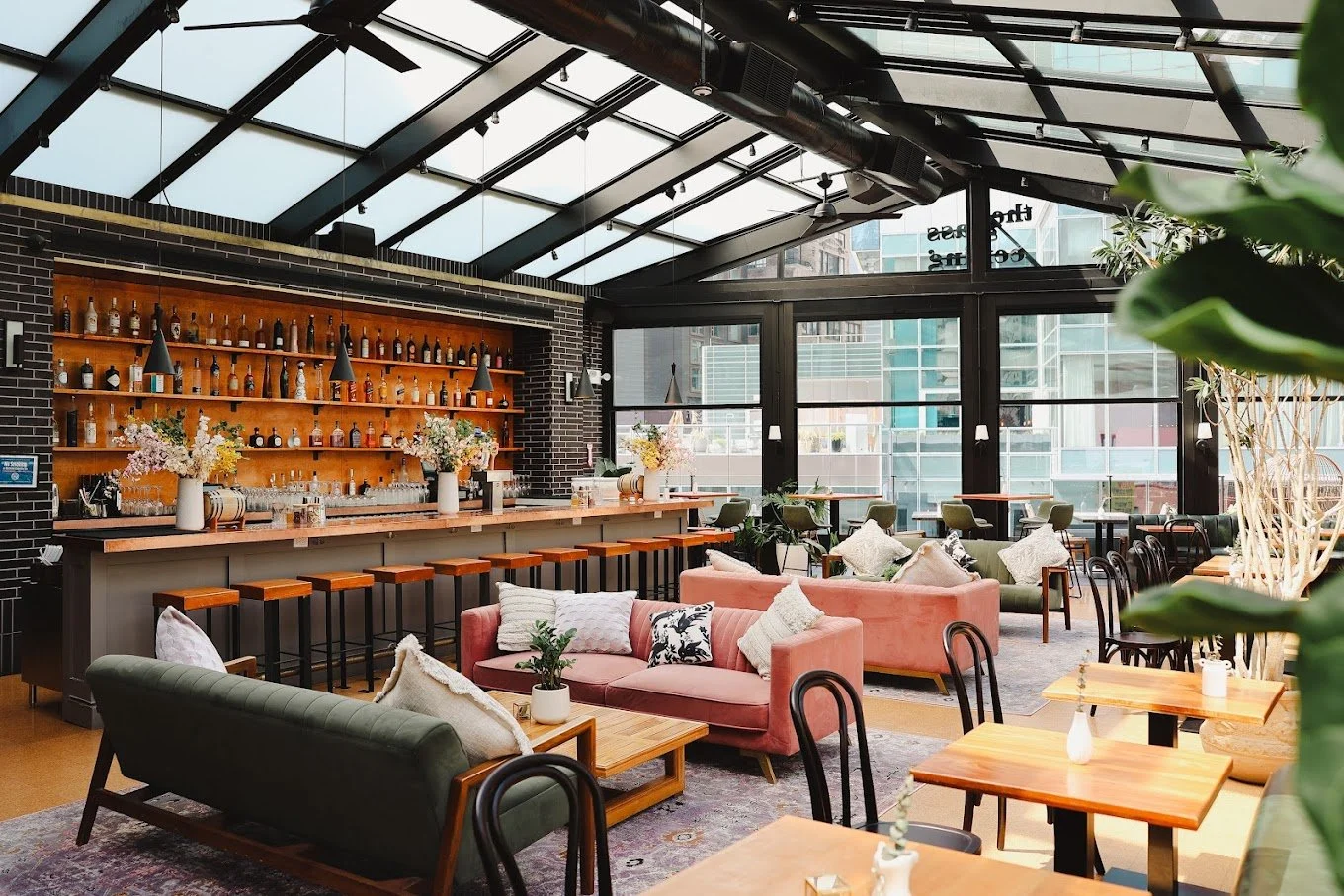 AMERICAN ROOFTOP BAR & GRILL - Interior of a modern, stylish restaurant or cafe with a bar area, seating arrangements including sofas and chairs, large windows, and decorative plants.