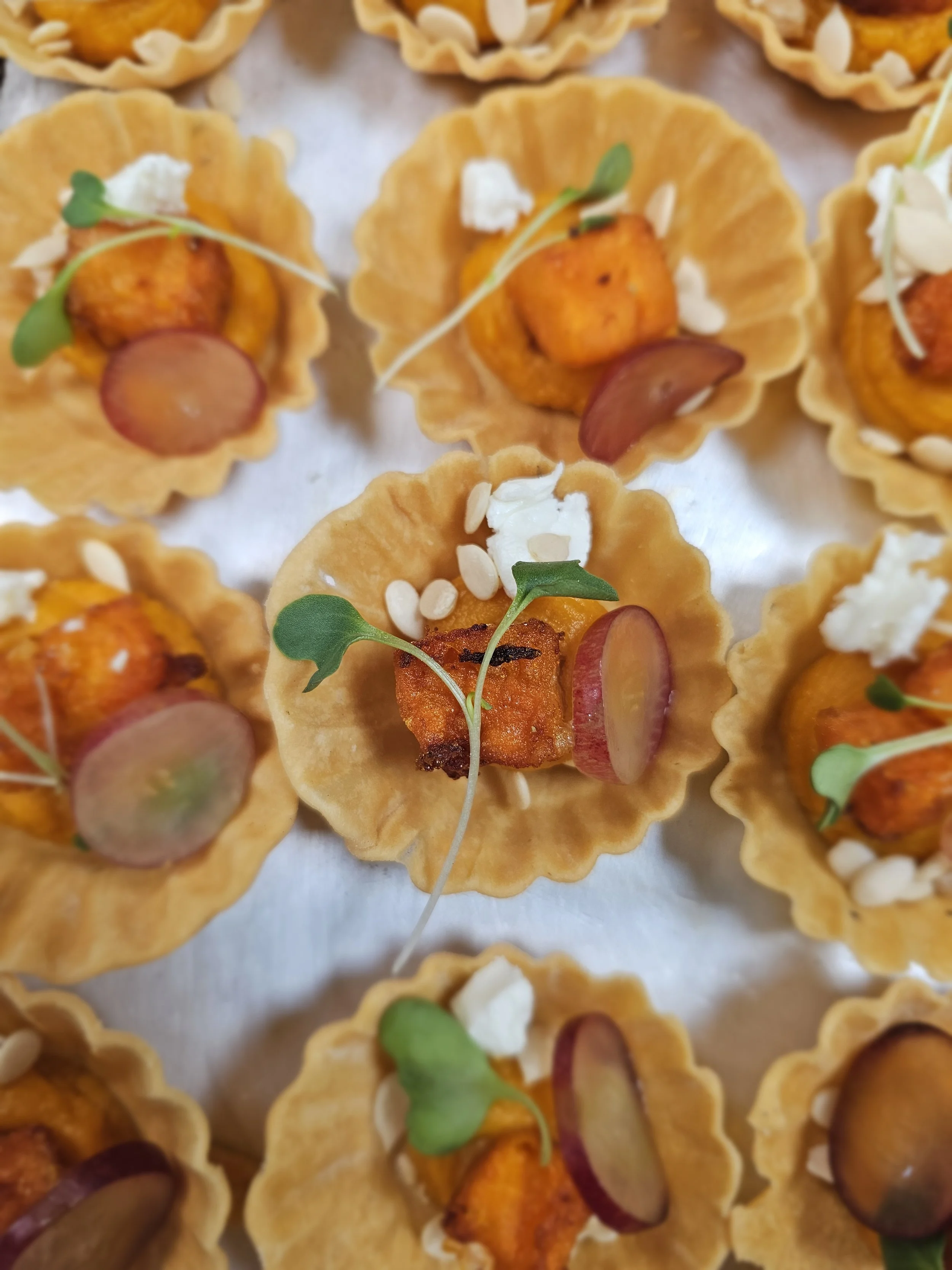 Miniature savory tartlets filled with roasted butternut squash, garnished with microgreens, sliced radish, and cheese.