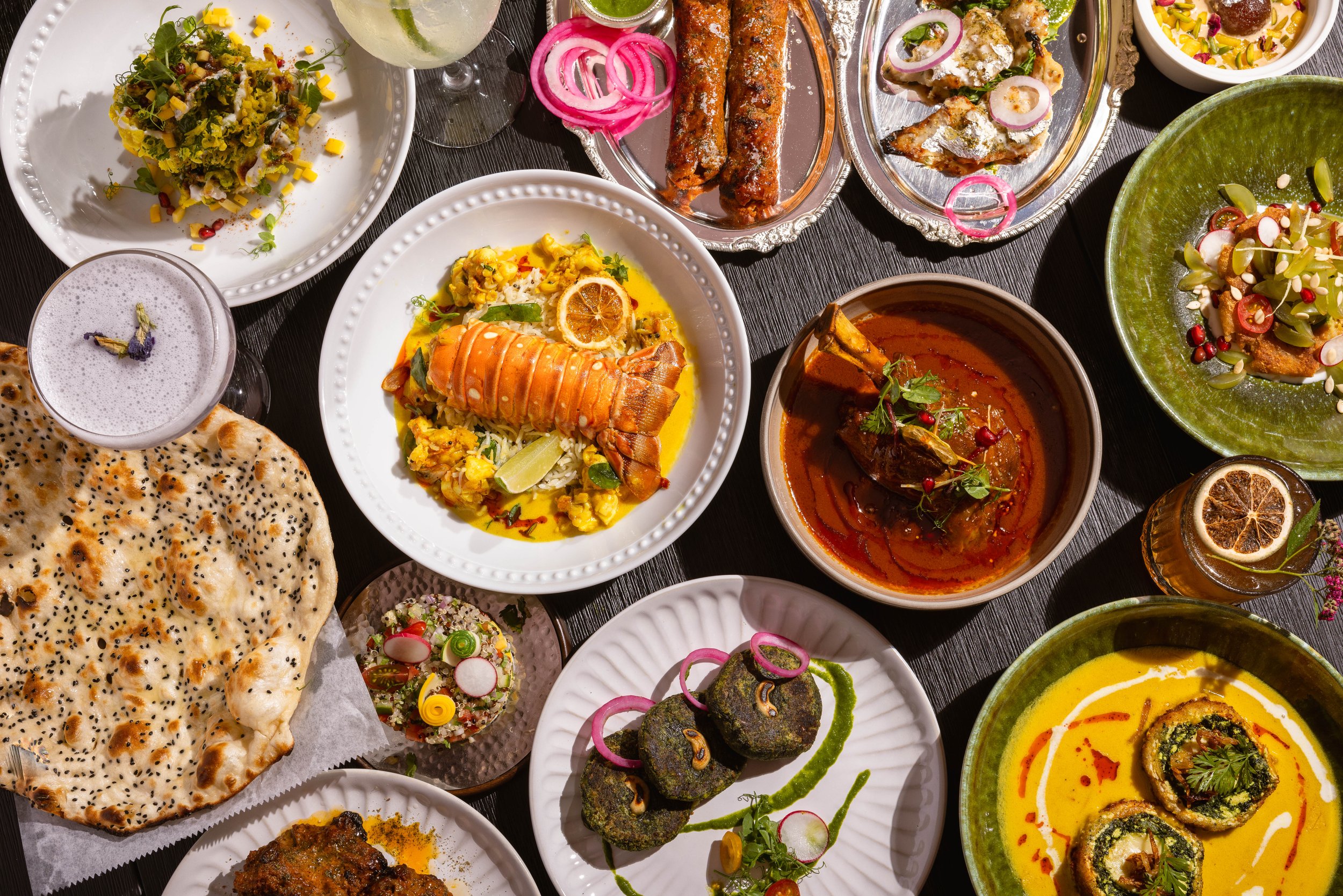A variety of Indian dishes on a table, including naan bread, curries, grilled meats, rice dishes, and garnished vegetables, with drinks and decorative garnishes. Made at Baazi.