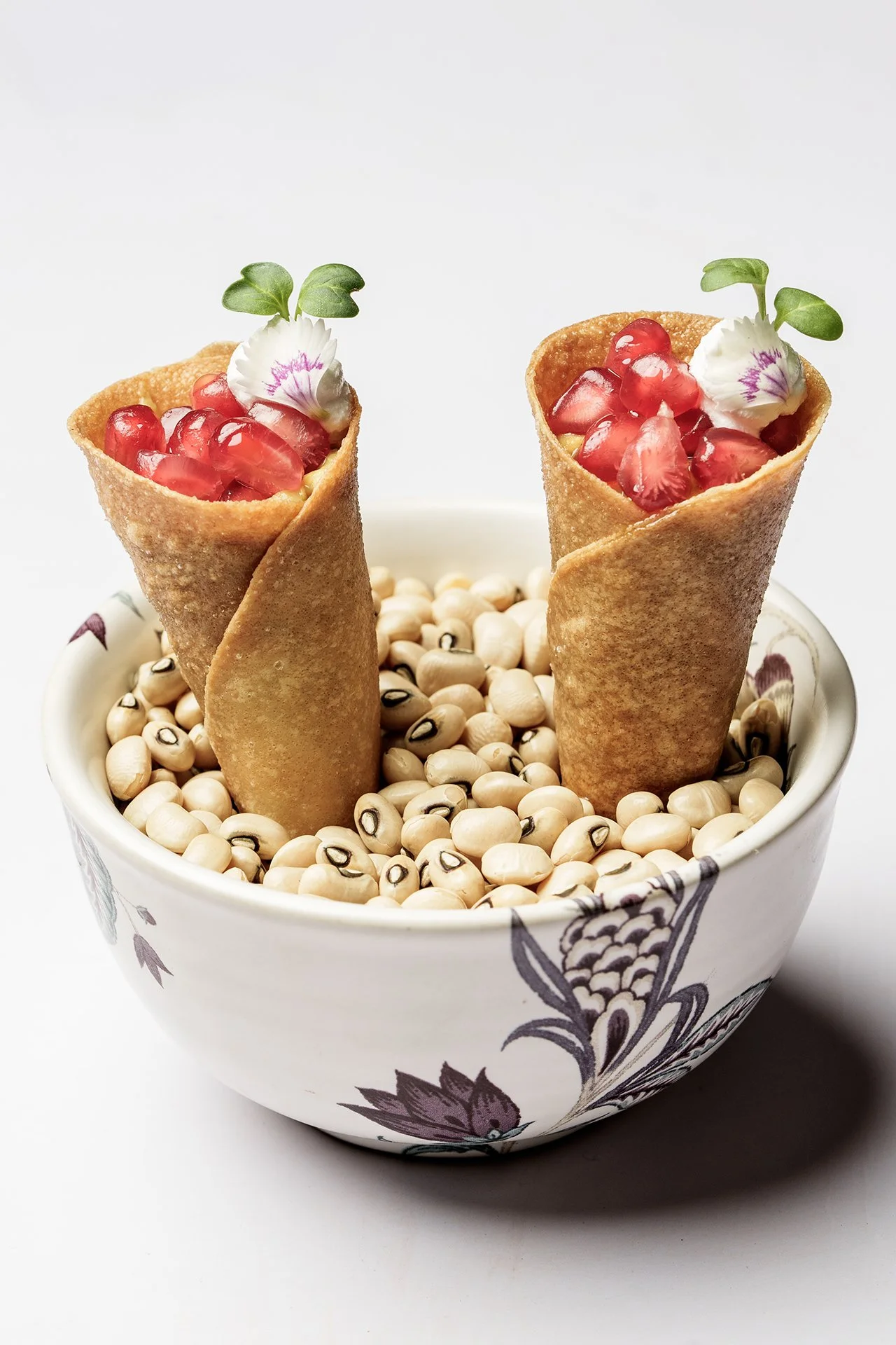 Two dessert cones filled with diced strawberries, garnished with edible flowers and microgreens, in a bowl filled with white beans.