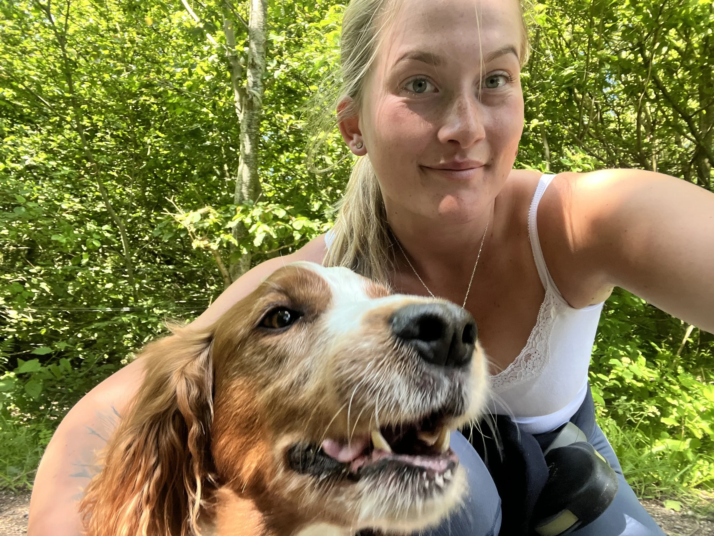 A woman with blonde hair and a white tank top taking a selfie with her dog in a forested area.