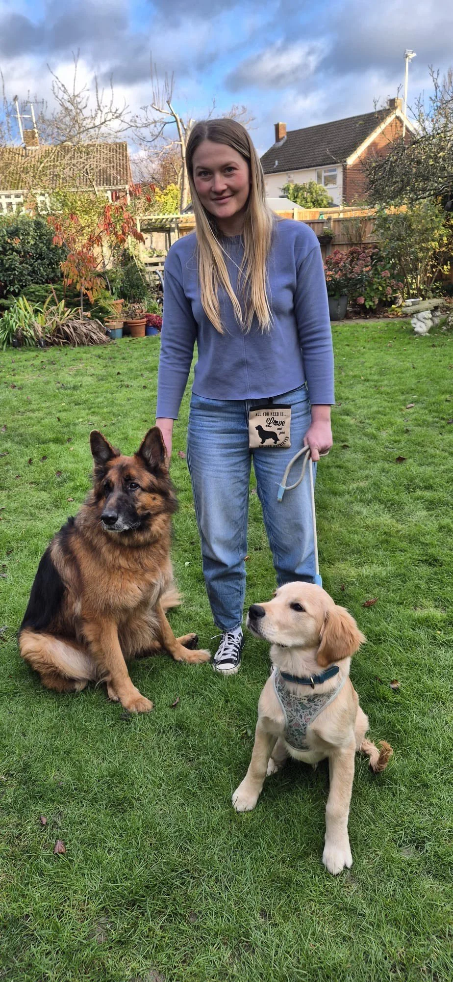 A young woman with long blonde hair standing on a grassy backyard, smiling, holding the leashes of a German Shepherd and a golden retriever puppy, with trees, bushes, and houses in the background on a partly cloudy day.