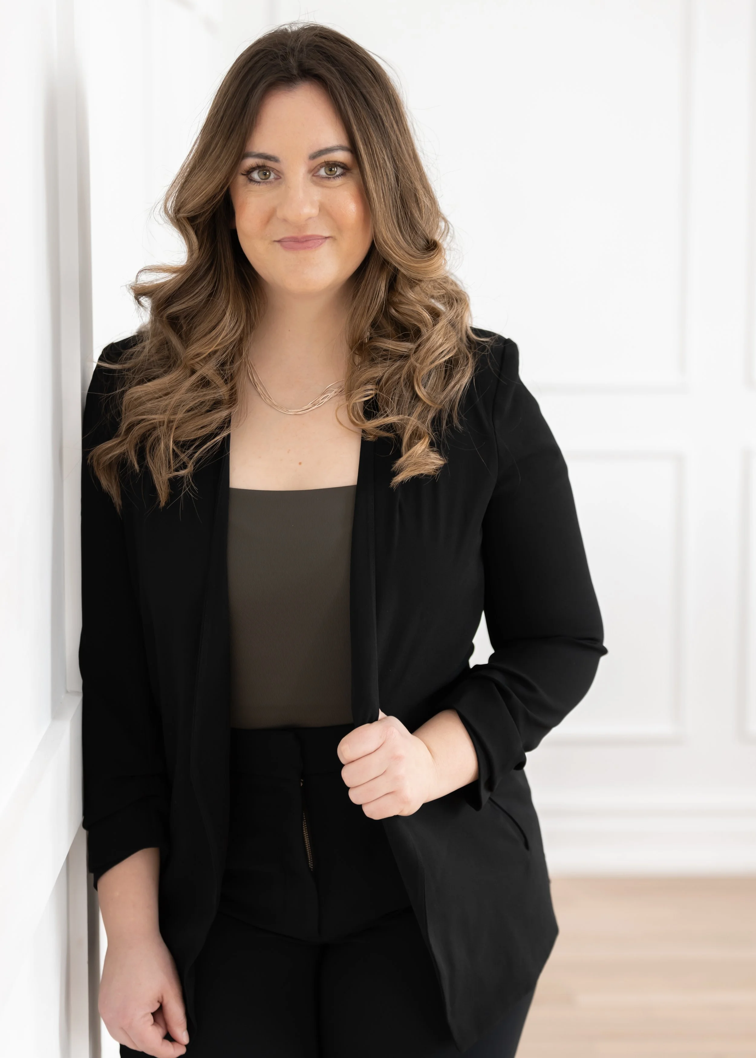 A woman with long wavy hair wearing a black blazer and dark pants, standing indoors against a white paneled wall.