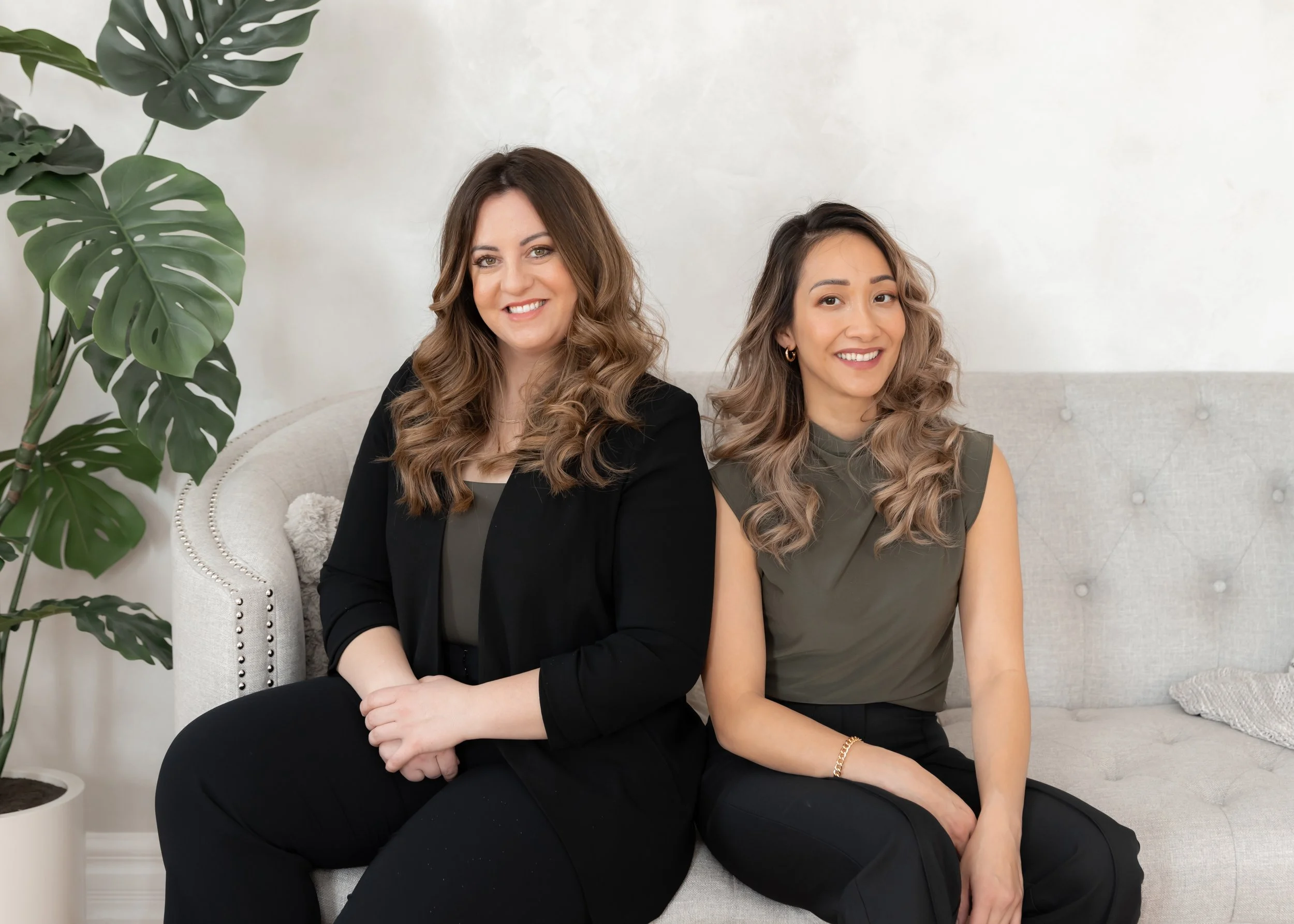 Two women sitting on a light-colored sofa in a room with a large green plant next to them. Both women are smiling and have wavy, shoulder-length hair. The woman on the left is wearing a black blazer and black pants, while the woman on the right is wearing a sleeveless olive-green top and black pants.