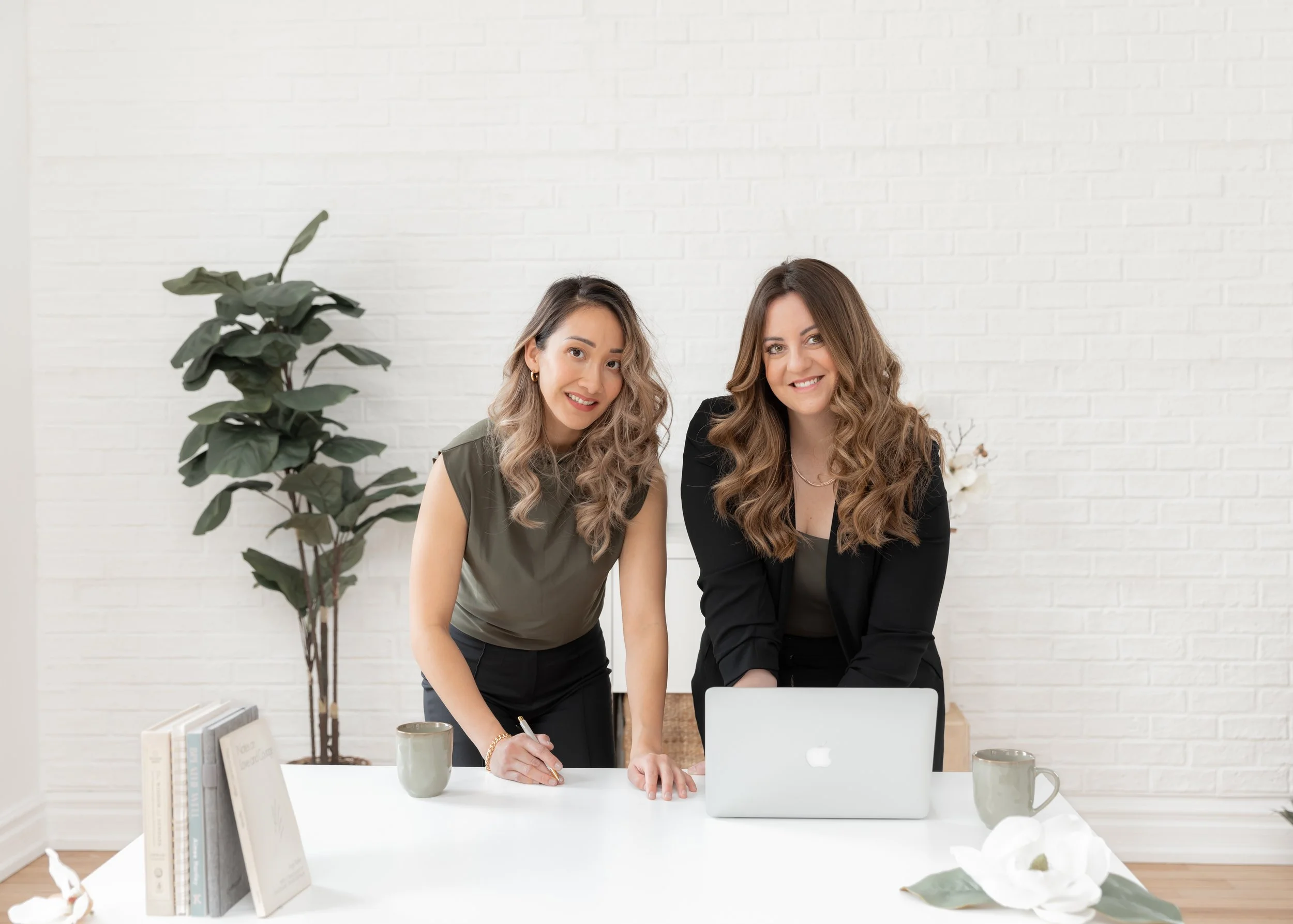 Two women in an office setting, smiling at the camera, one standing and one leaning on a table, with a laptop, books, potted plant, and mugs on the table, and a white brick wall in the background.