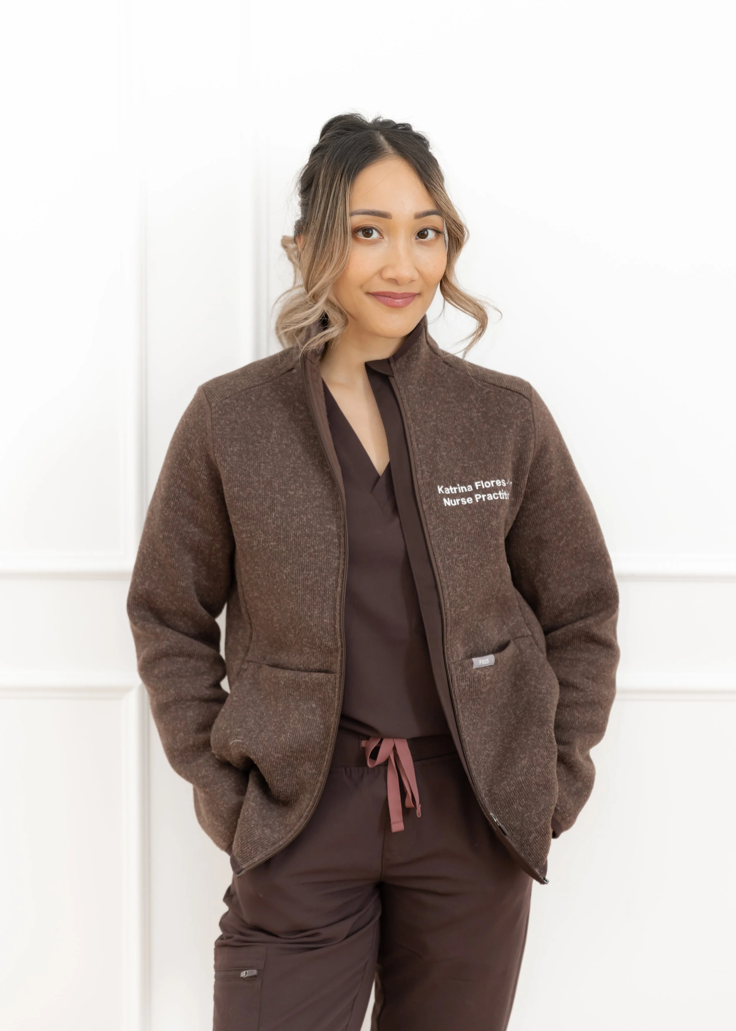 A woman in brown scrubs and a brown jacket standing in front of a white wall.
