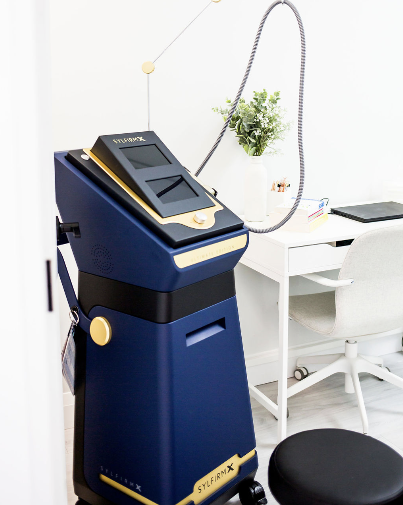 A medical or aesthetic treatment machine with a screen and a flexible arm, placed next to a white desk with a potted plant, books, and a laptop, in a bright, modern office or clinic room.