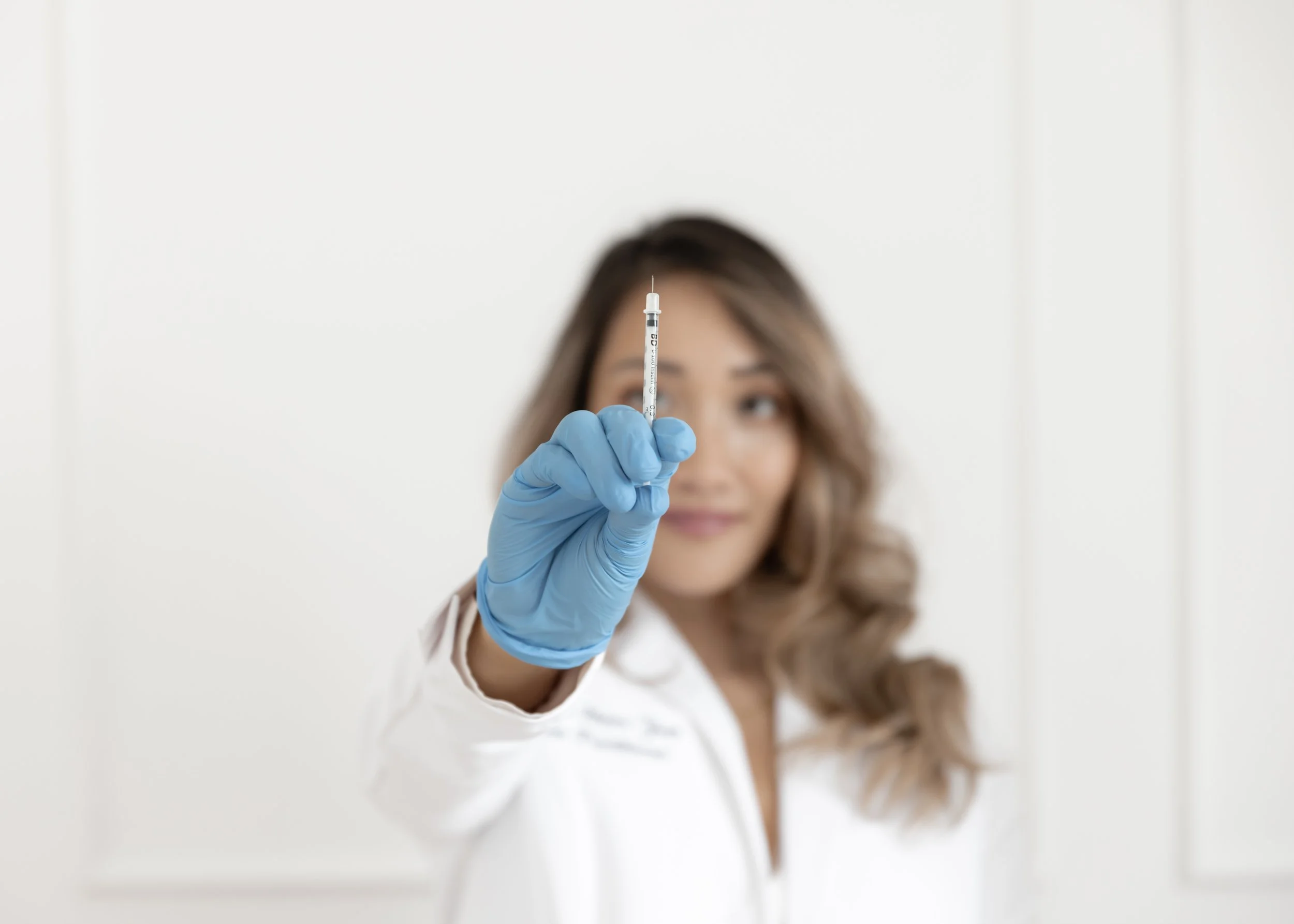 A woman wearing a white coat and blue gloves holding up a syringe, with a blurred background.