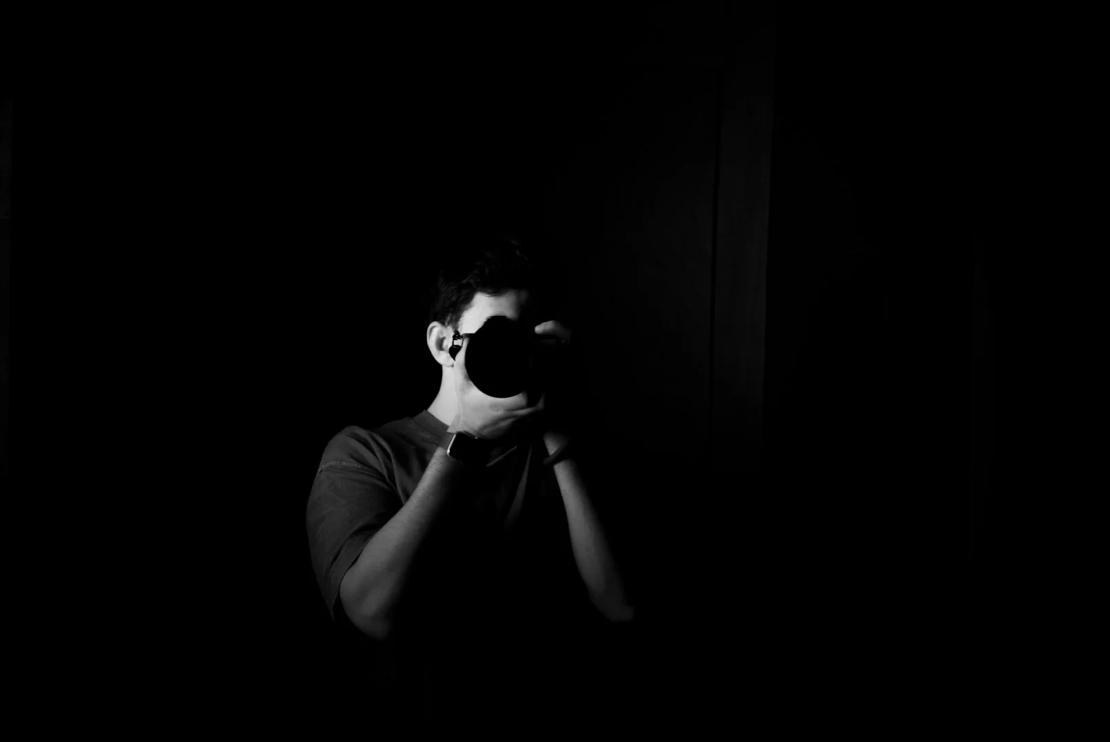 A person taking a mirror selfie with a camera in a dark room, dressed in casual clothing, with a smartwatch on wrist, black and white photography.