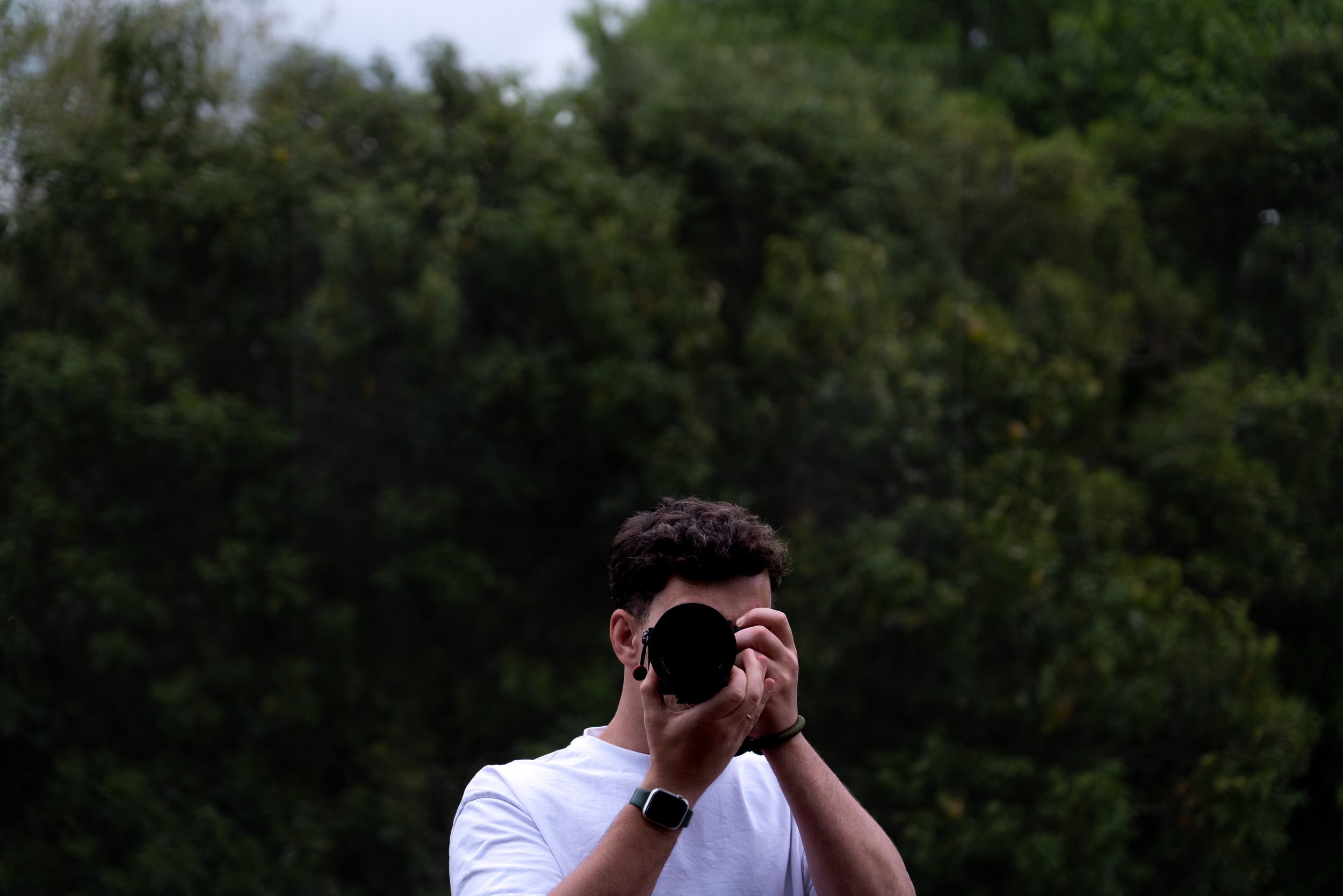 Person with short curly hair, wearing a white t-shirt, holding a camera up to their face outdoors with green trees in the background.