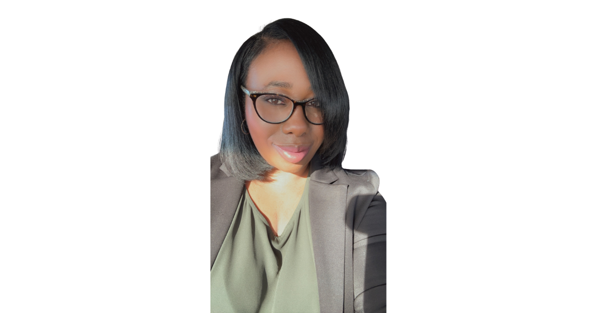 A woman with shoulder-length black hair, wearing glasses, a dark blazer, and a green top, taking a selfie against a white background.