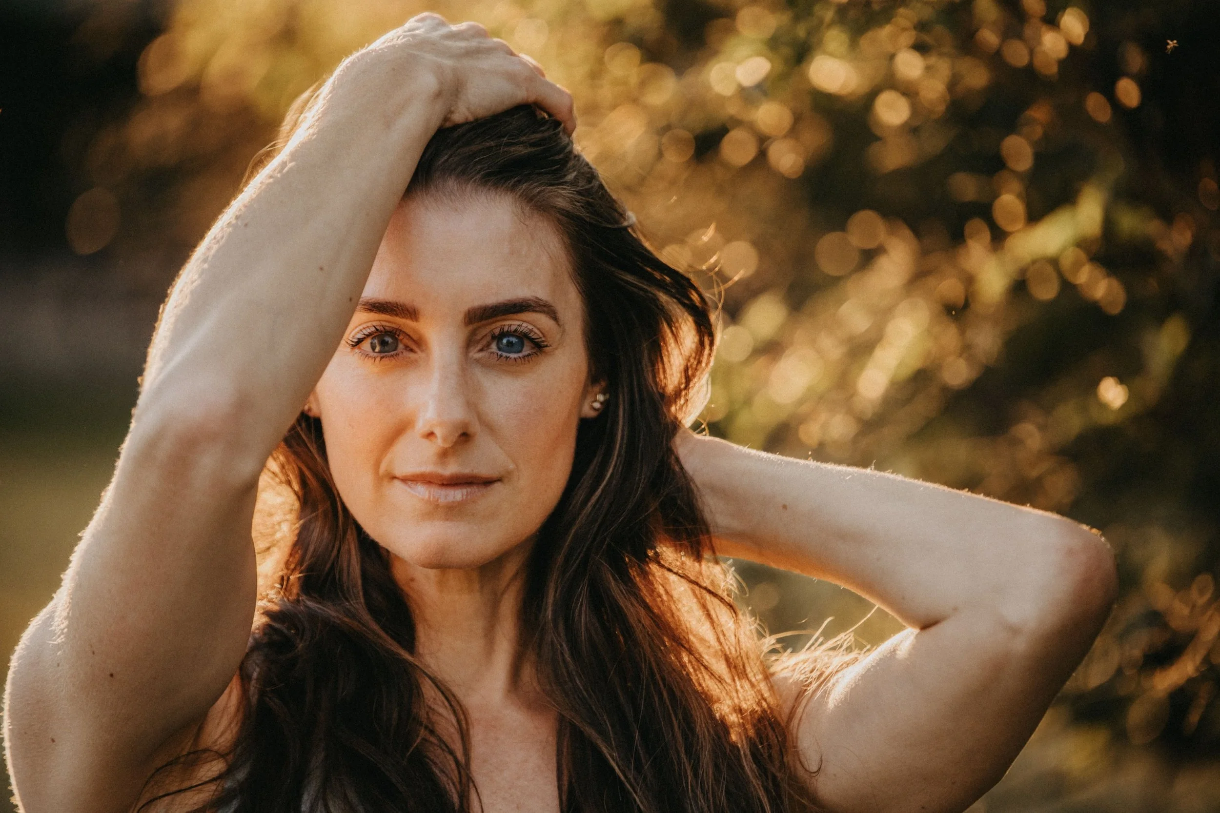 A woman with long brown hair and blue eyes looking at the camera during sunset, with her arms raised and hands resting on her head, outdoors with a blurred background of trees and bokeh lights.