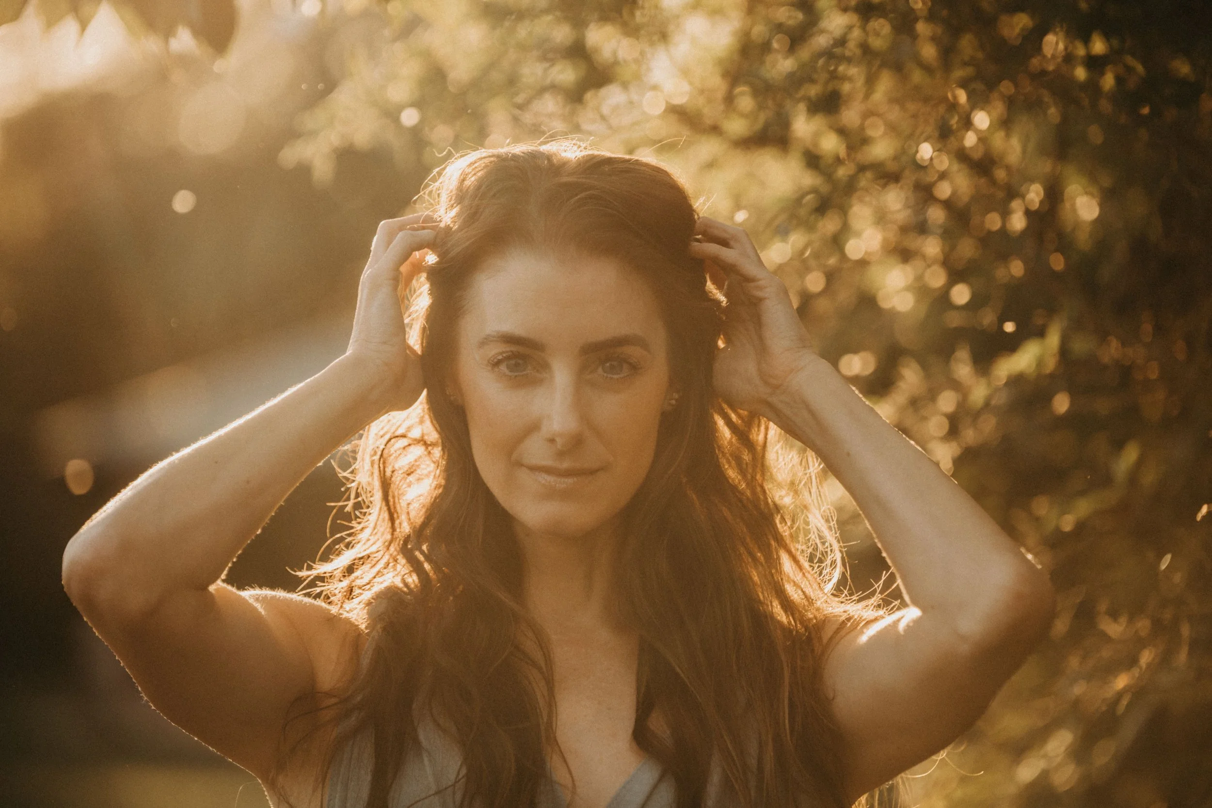 a woman with red hair outdoors during sunset, holding her hair back, looking directly at the camera.