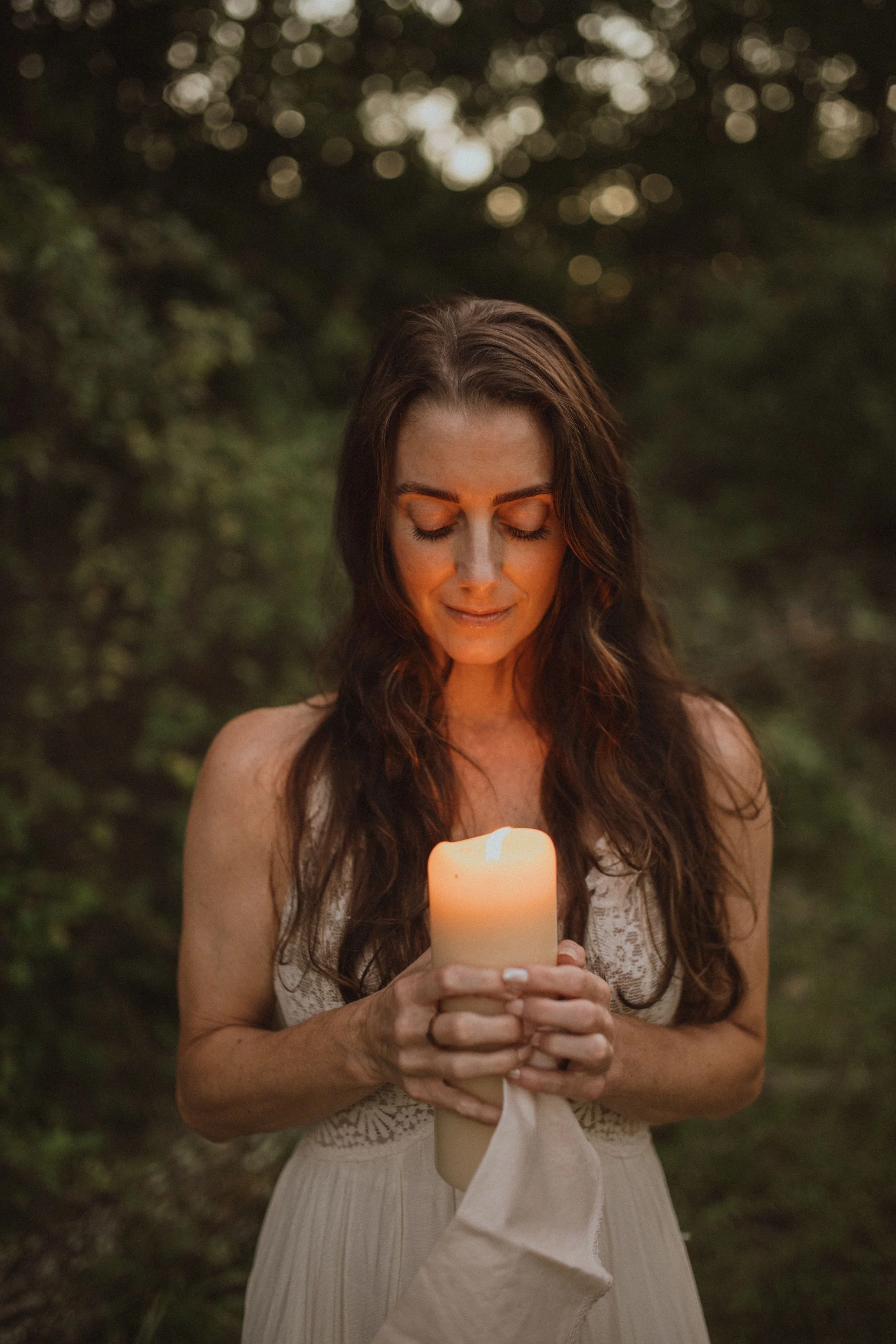 A woman with long brown hair smiling with eyes closed, holding a lit white candle with a cloth wrapped around its base, standing outdoors in a wooded area at dusk.