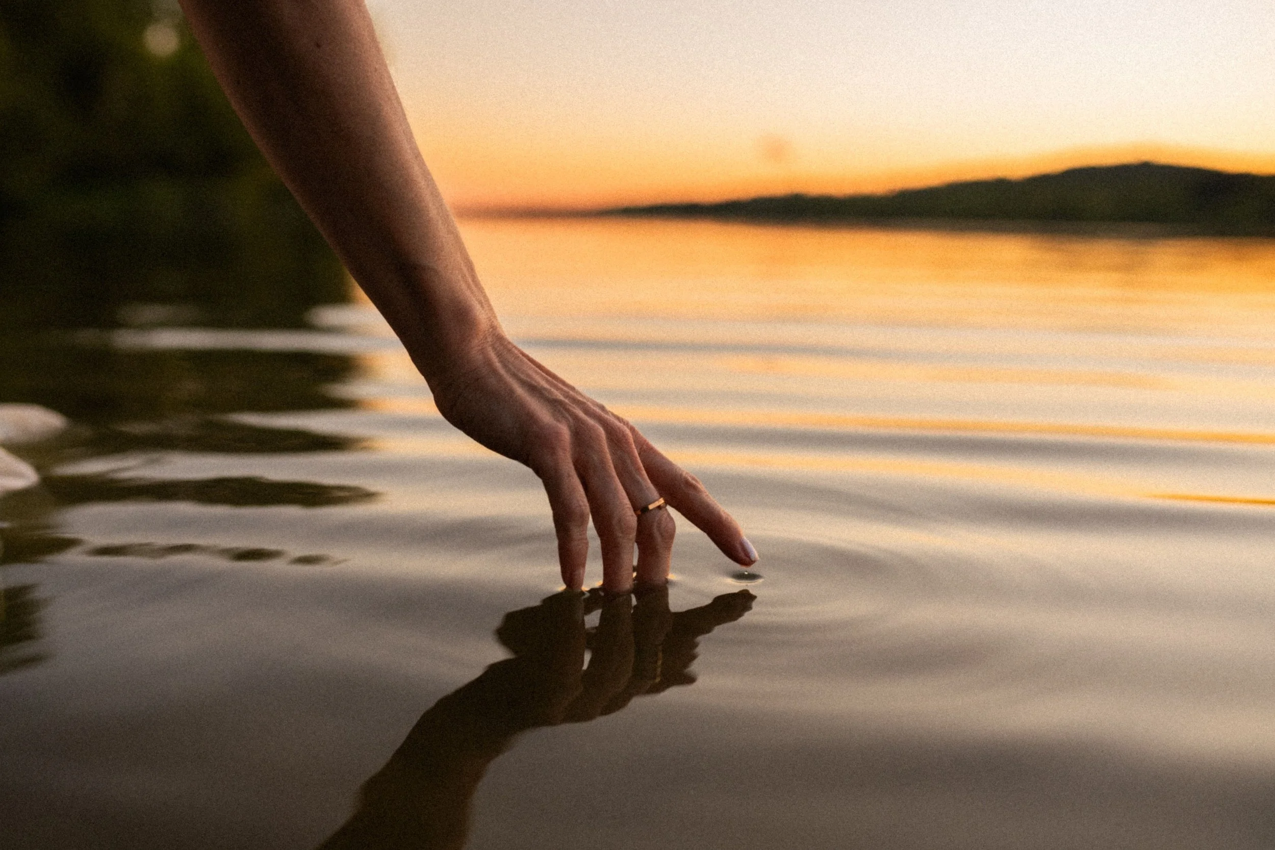 Close-up of a person's hand reaching into a calm body of water at sunset, with orange and pink hues in the sky and distant hills in the background.