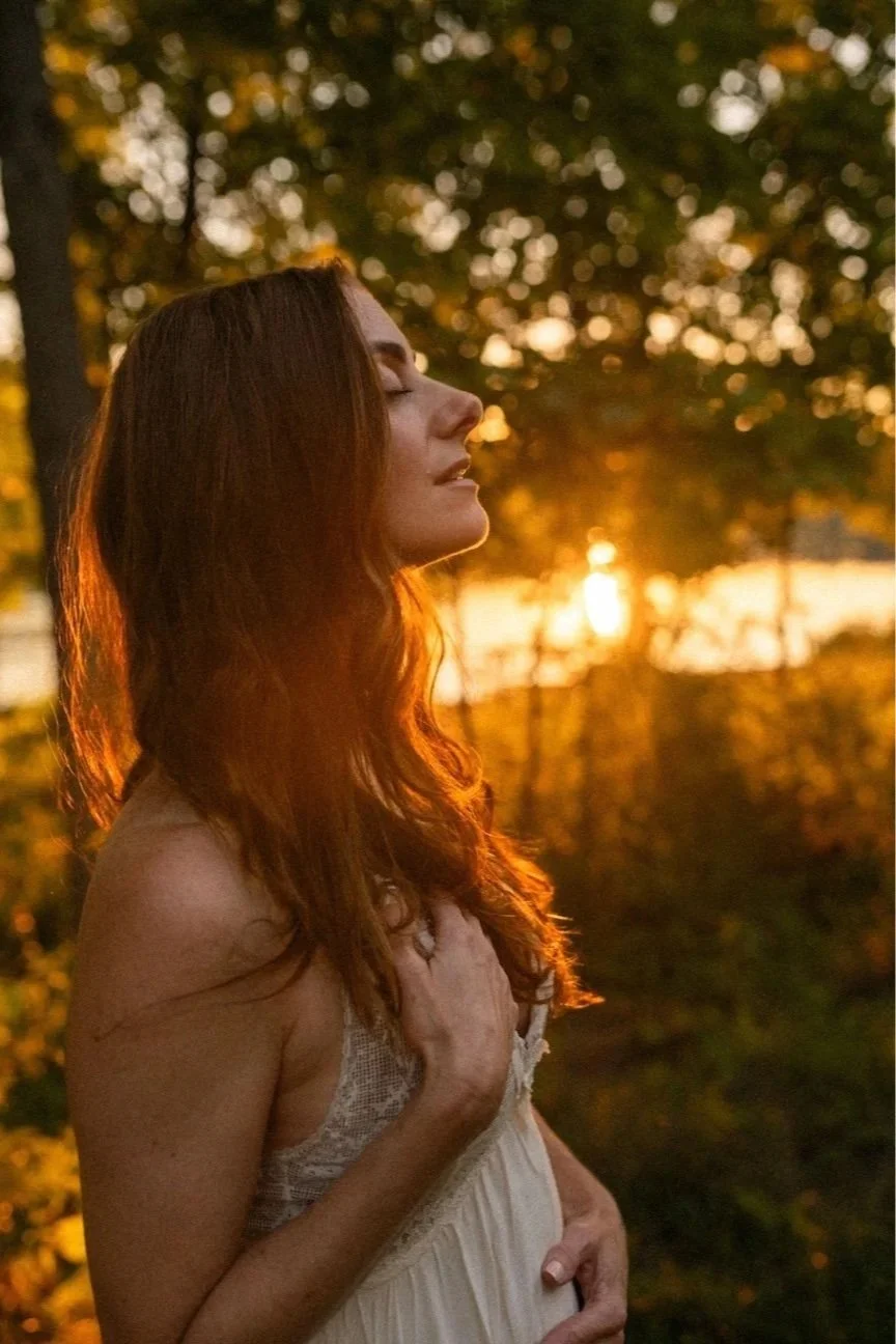A pregnant woman with long red hair standing outdoors during sunset, with eyes closed and hand resting on her belly.