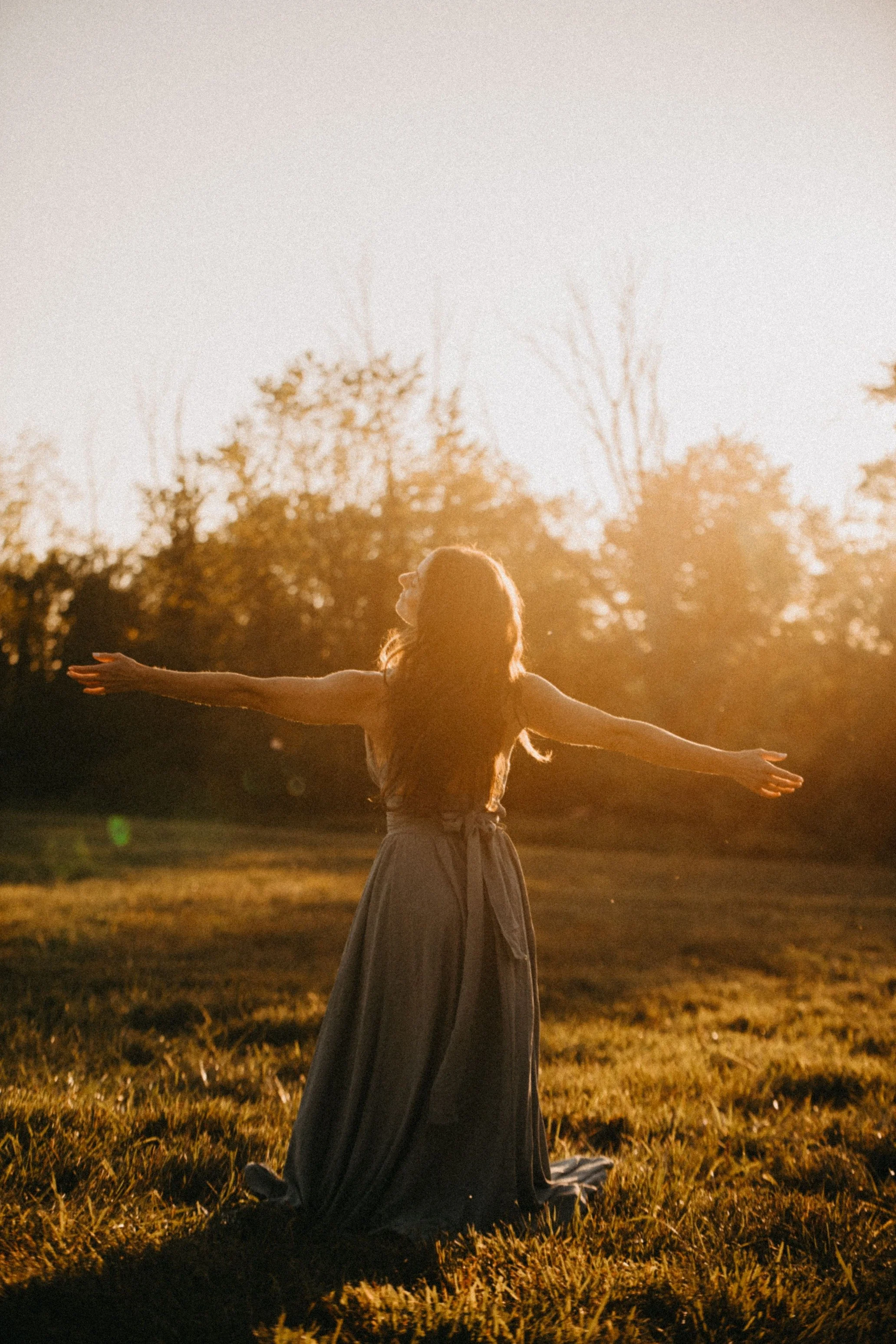 A woman with long hair wearing a flowing dress is standing outdoors in a grassy field at sunset with her arms outstretched and her face turned upward.