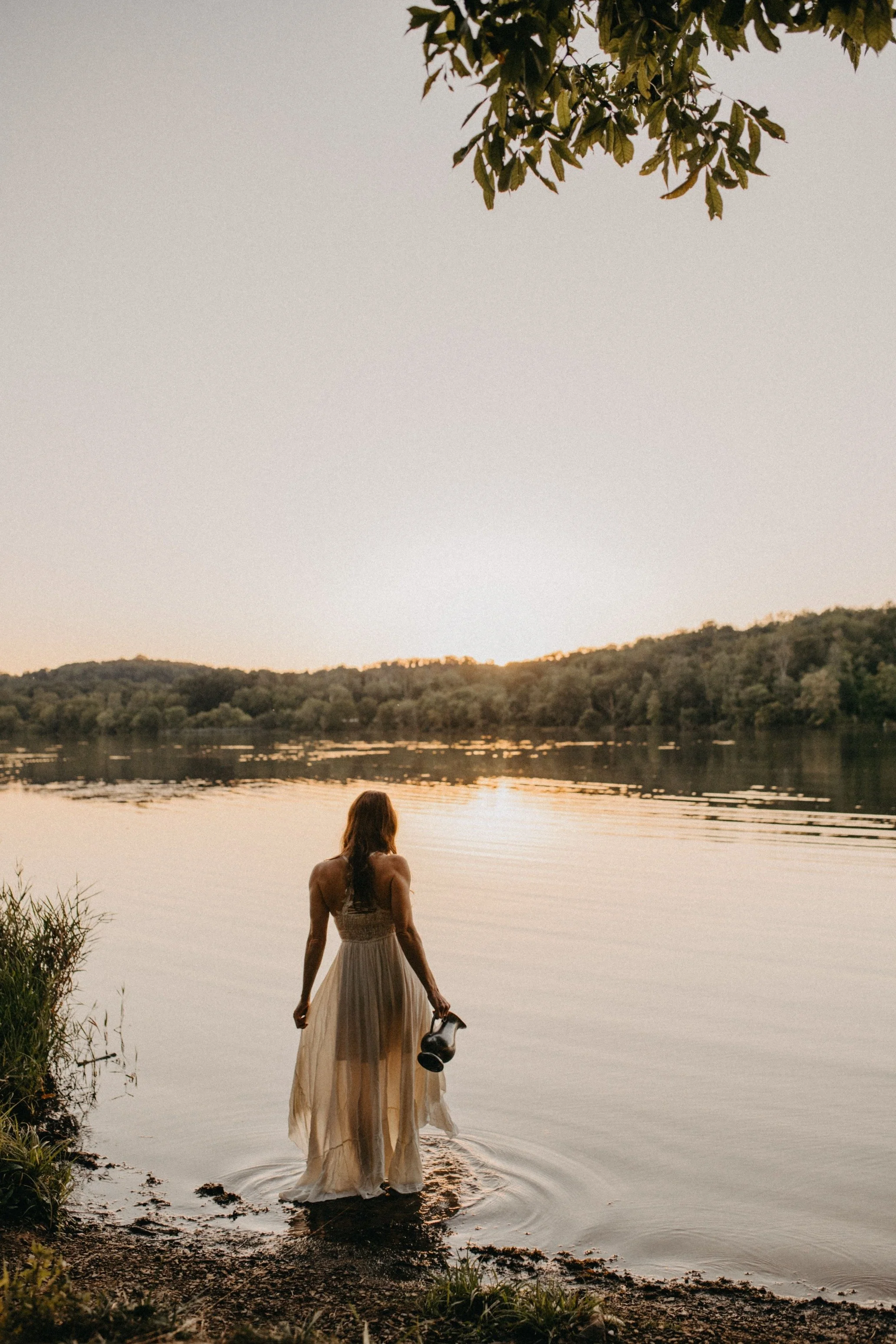 A woman in a flowing white dress walking into a lake at sunset, holding a pair of shoes.