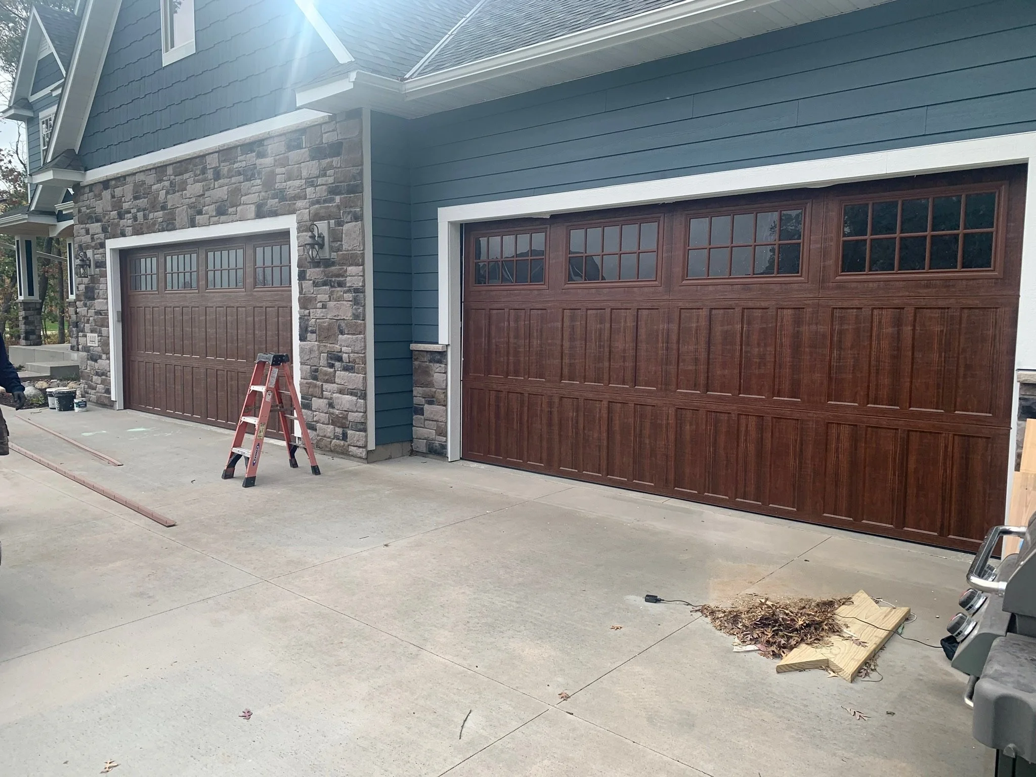 Construction progress on a two-car garage with wooden doors, blue siding, stone accents, and outdoor lighting fixtures, with construction tools and materials on the driveway.