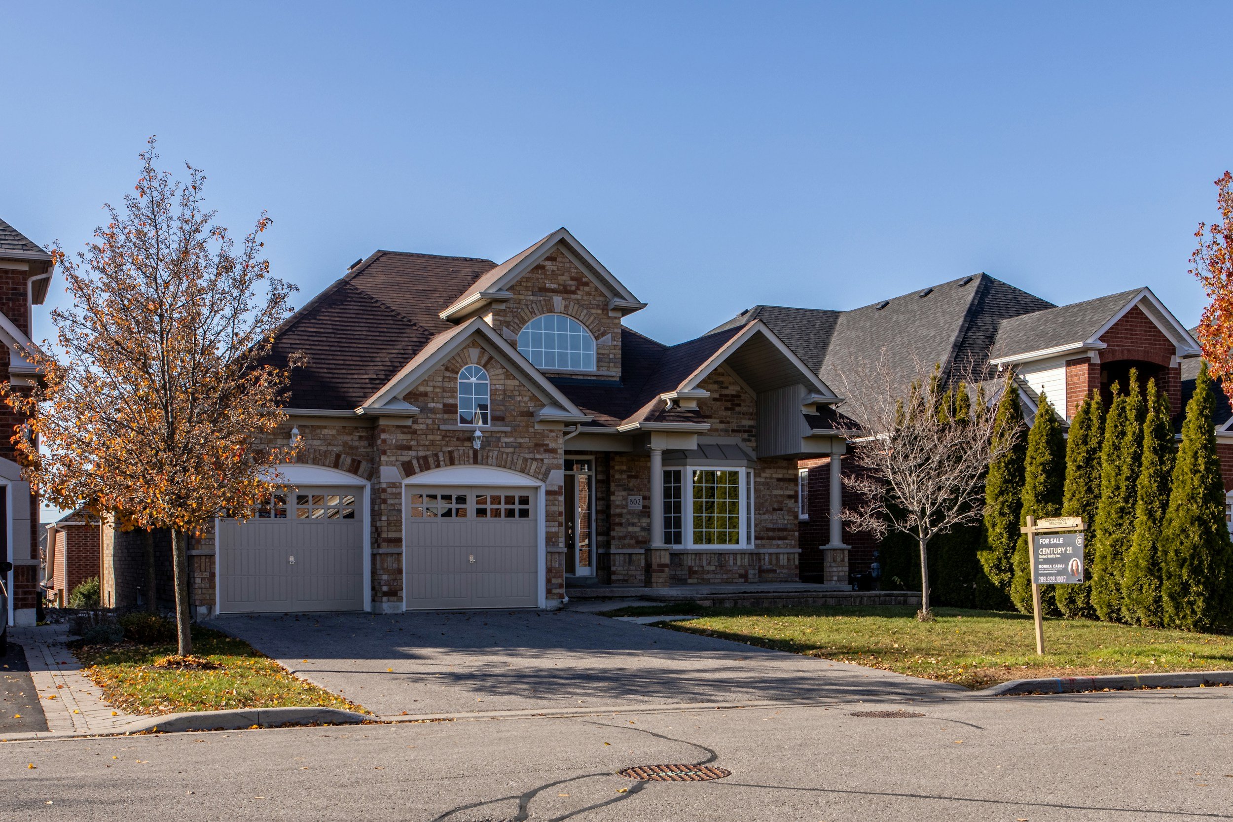 A two-story brick house with a three-car garage, a driveway, and a front yard with trees and a for sale sign.
