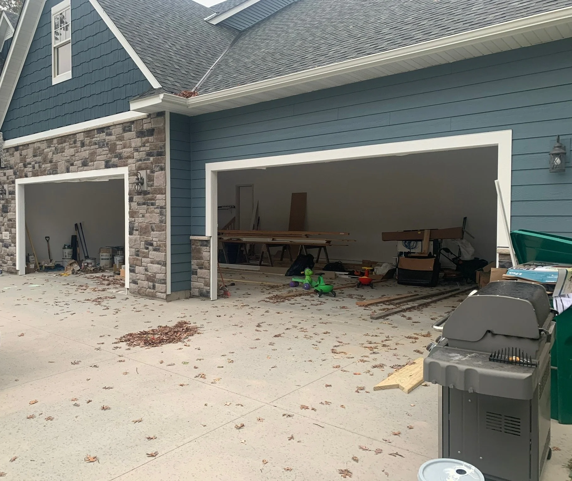 A house with two open garage bays, one with construction materials and tools inside, and the other with scattered toys and construction debris. The driveway has fallen leaves and a barbecue grill is on the right side.
