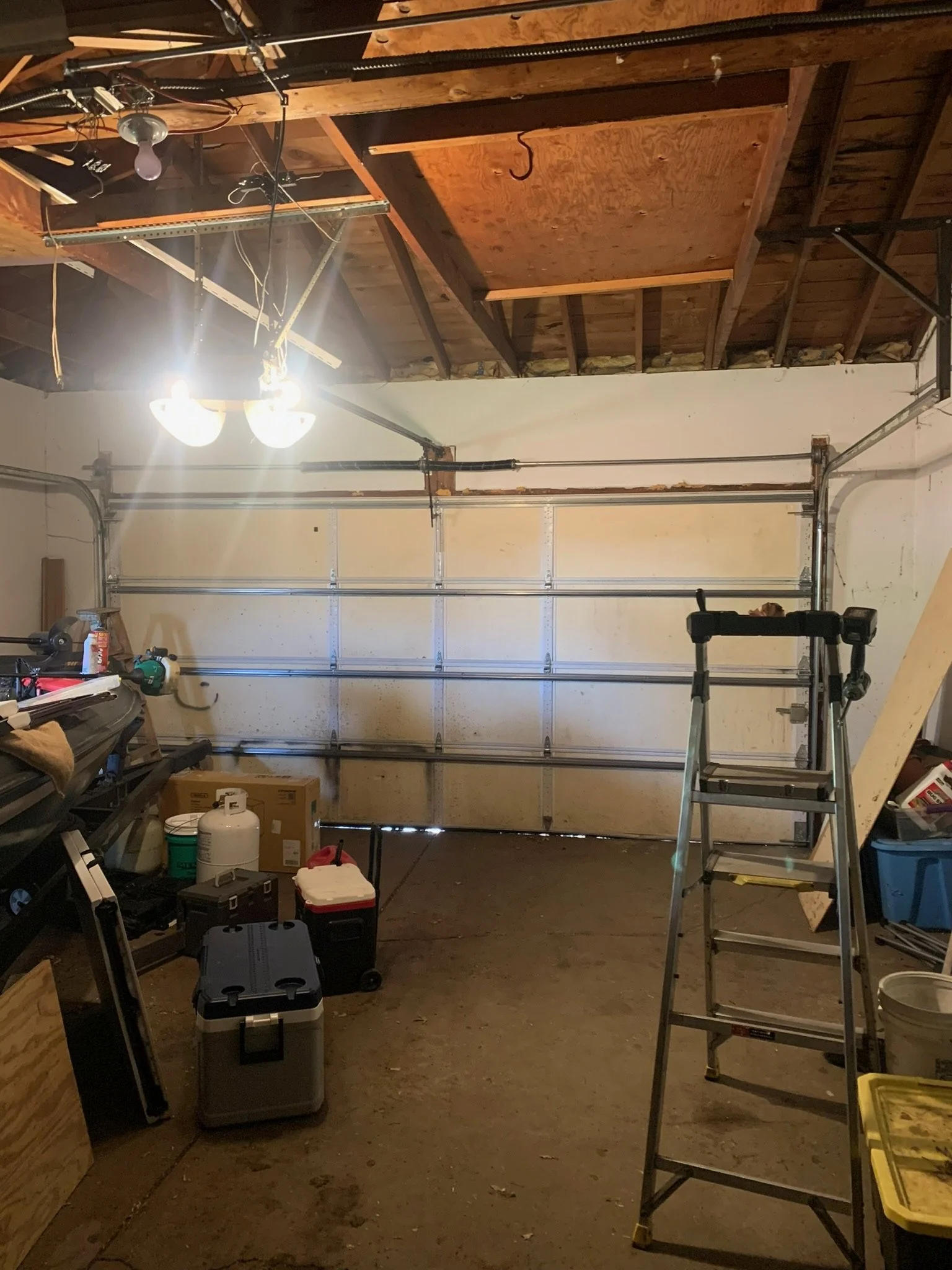An unfinished garage with a closed white garage door, a step ladder, and various tools and storage boxes around.