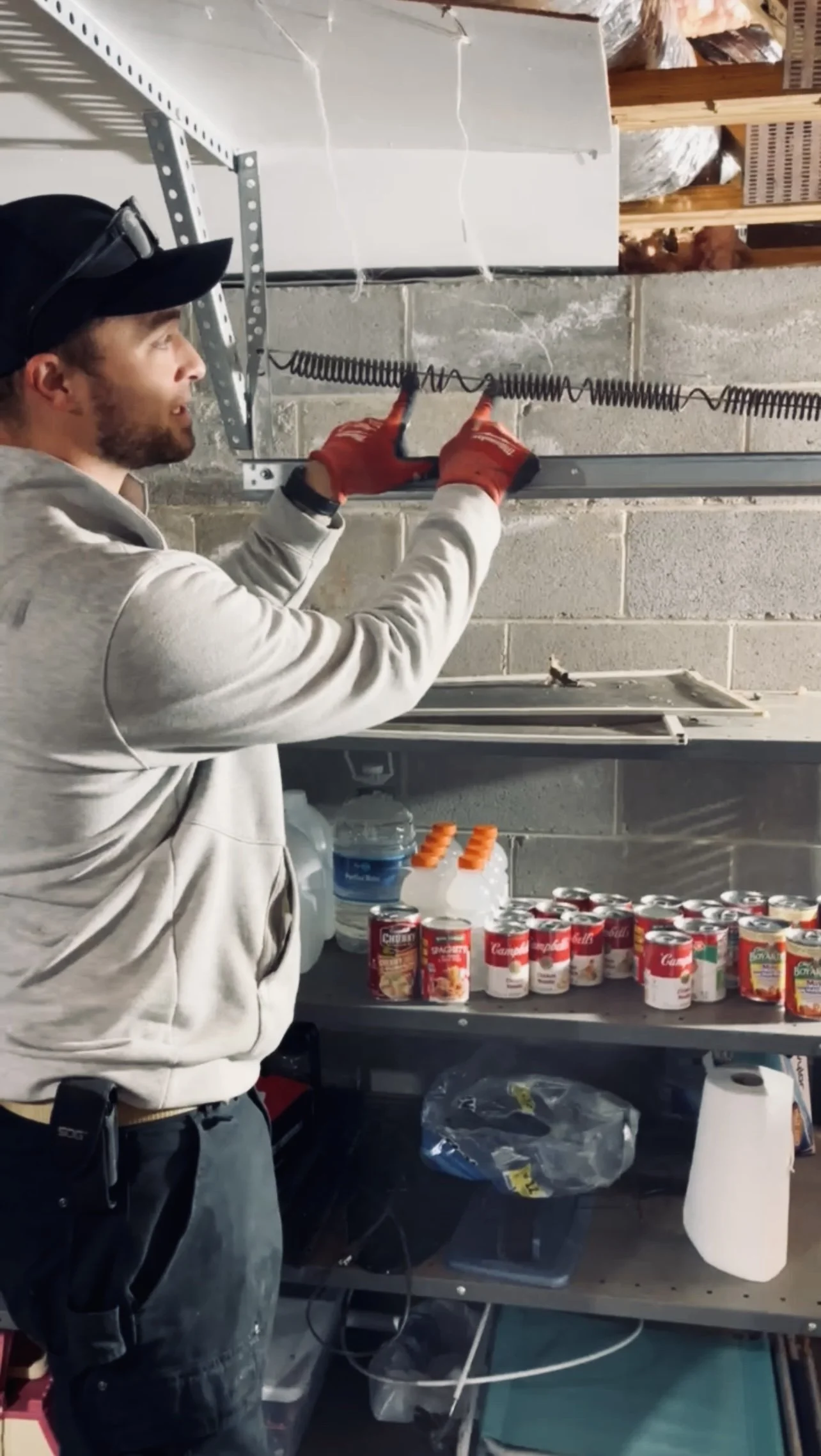 A man with a beard, wearing a gray hoodie, black cap, and red gloves, is working on electrical wiring in a garage or storage room. Shelves with canned goods, bottles, and cleaning supplies are visible in the background.