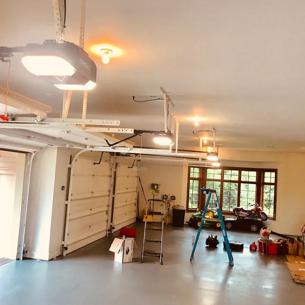 Garage with an open door, ceiling-mounted garage door openers, a ladder, and various tools and items stored near the back wall.
