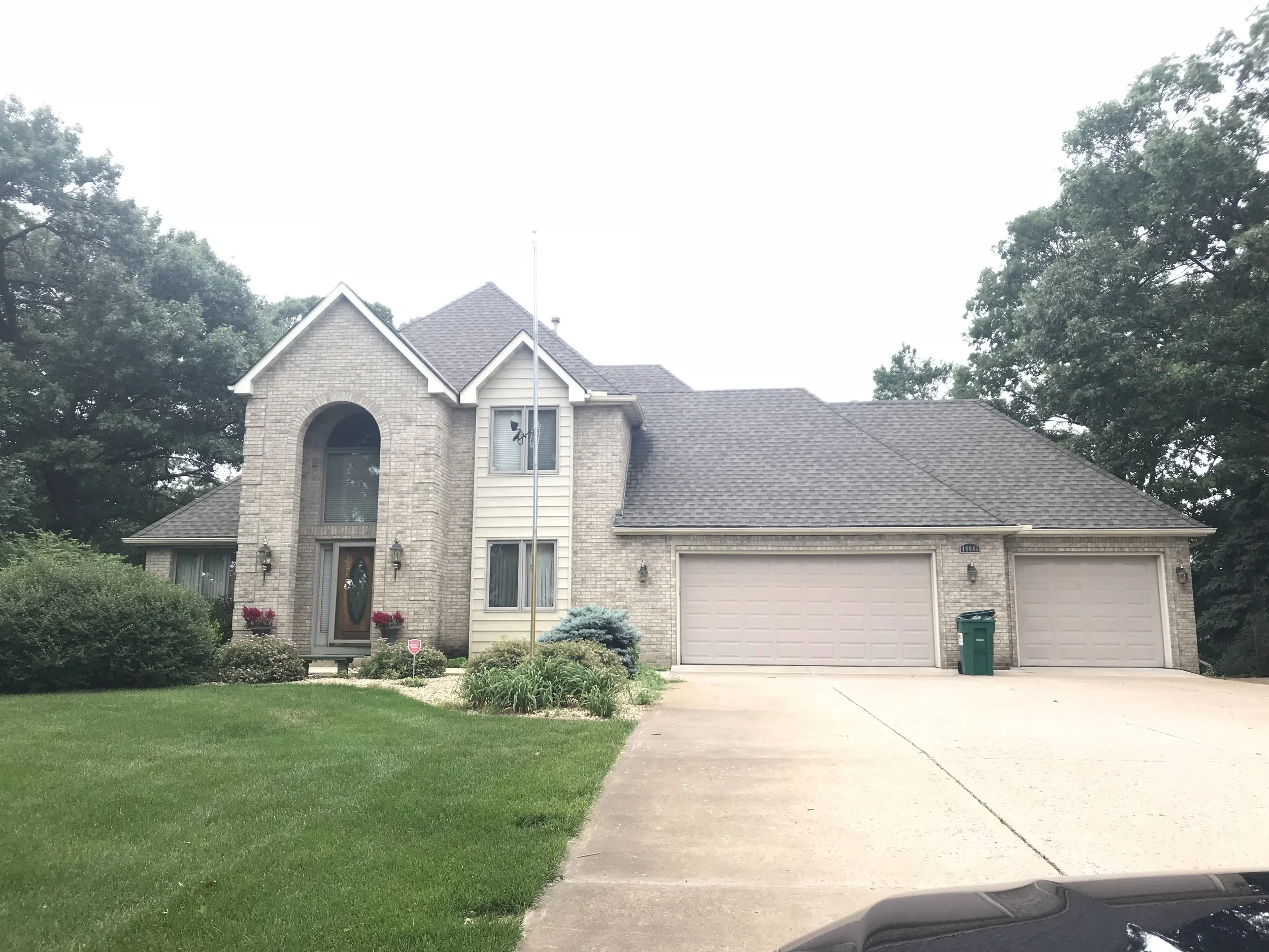 Front view of a two-story house with brick and siding, a three-car garage, a driveway, and green lawn with landscaped bushes and trees.