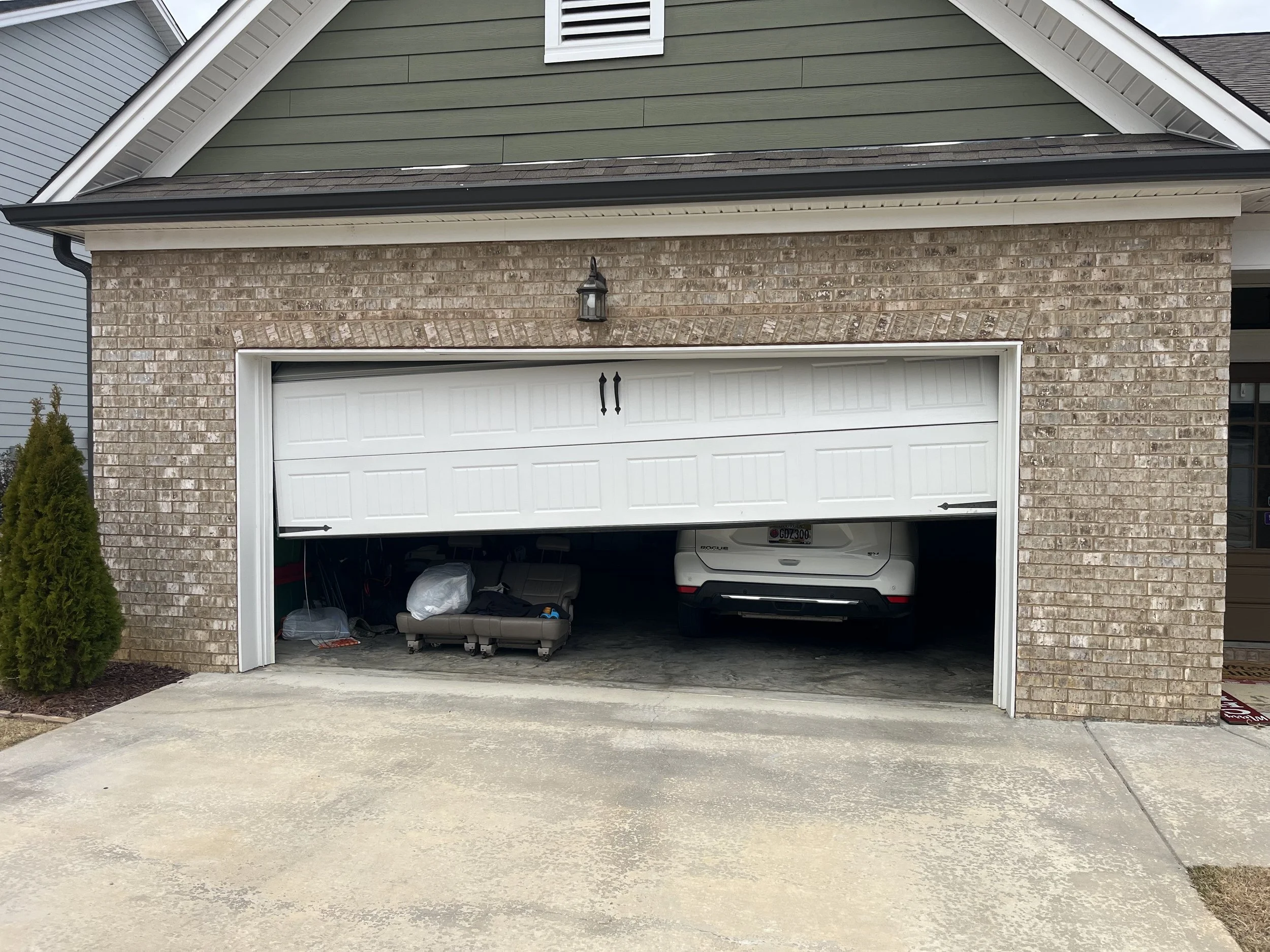 The image depicts a garage door that is partially open, with a white SUV parked inside. The garage has brick and siding exterior walls, with a small lamp above the door. The driveway is concrete, and there is a small bush to the left of the garage.