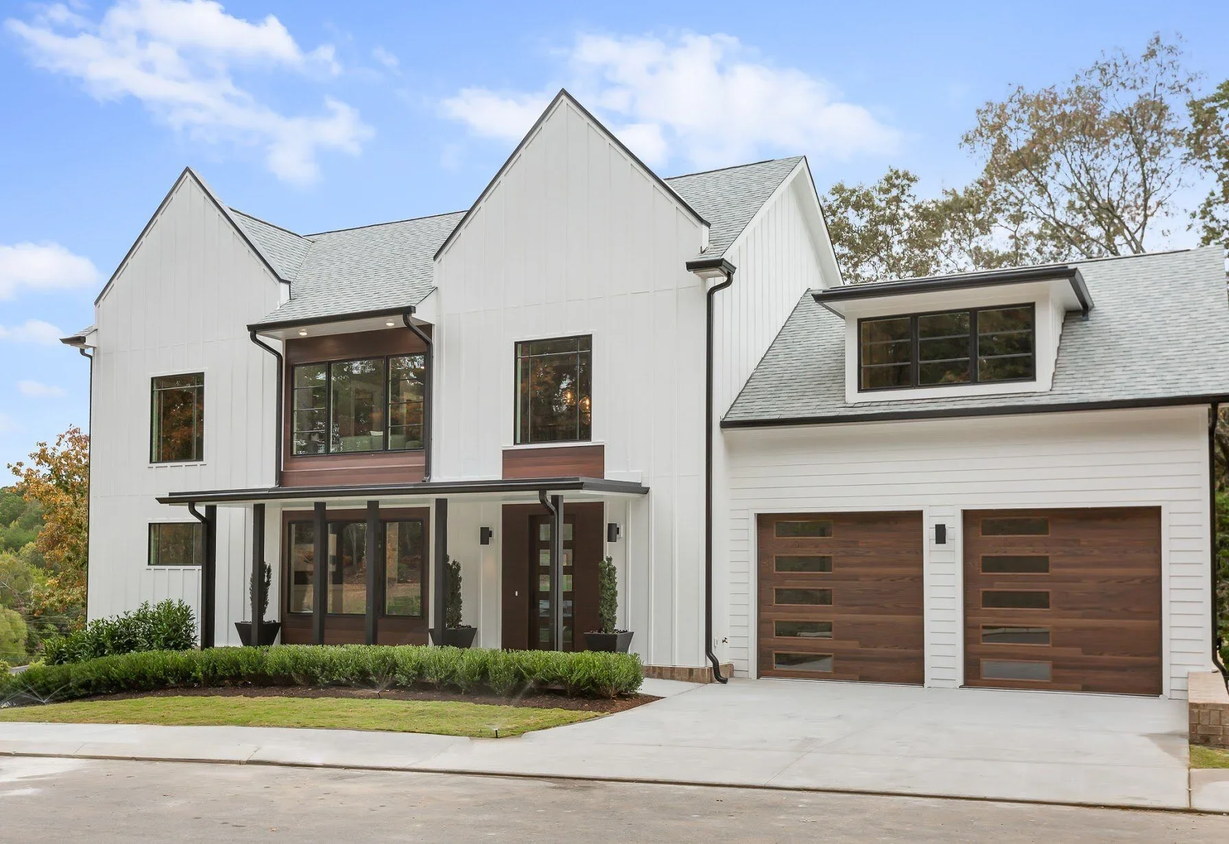 Modern white house with gray shingled roof, featuring large windows, a double garage with wooden doors, and a small front porch with black columns and potted plants, surrounded by greenery.