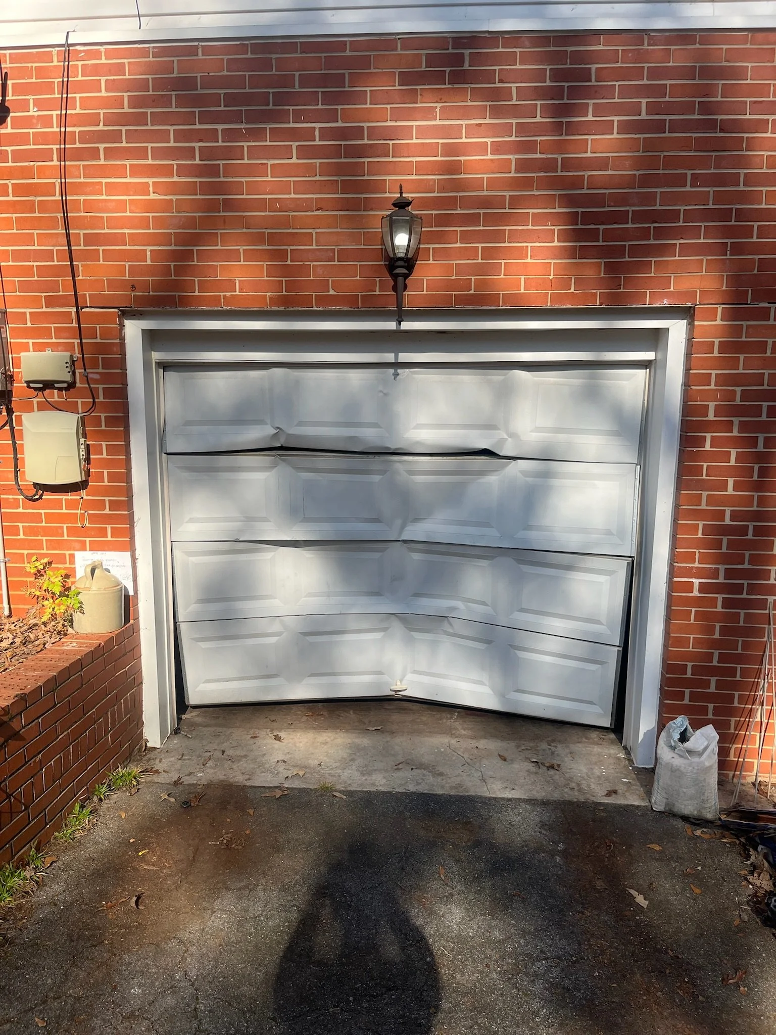 A white garage door on a brick house, with a black exterior lantern above it, and shadows cast on the door and house wall. There's a small garden bed on the left side and a bag on the right side of the driveway.