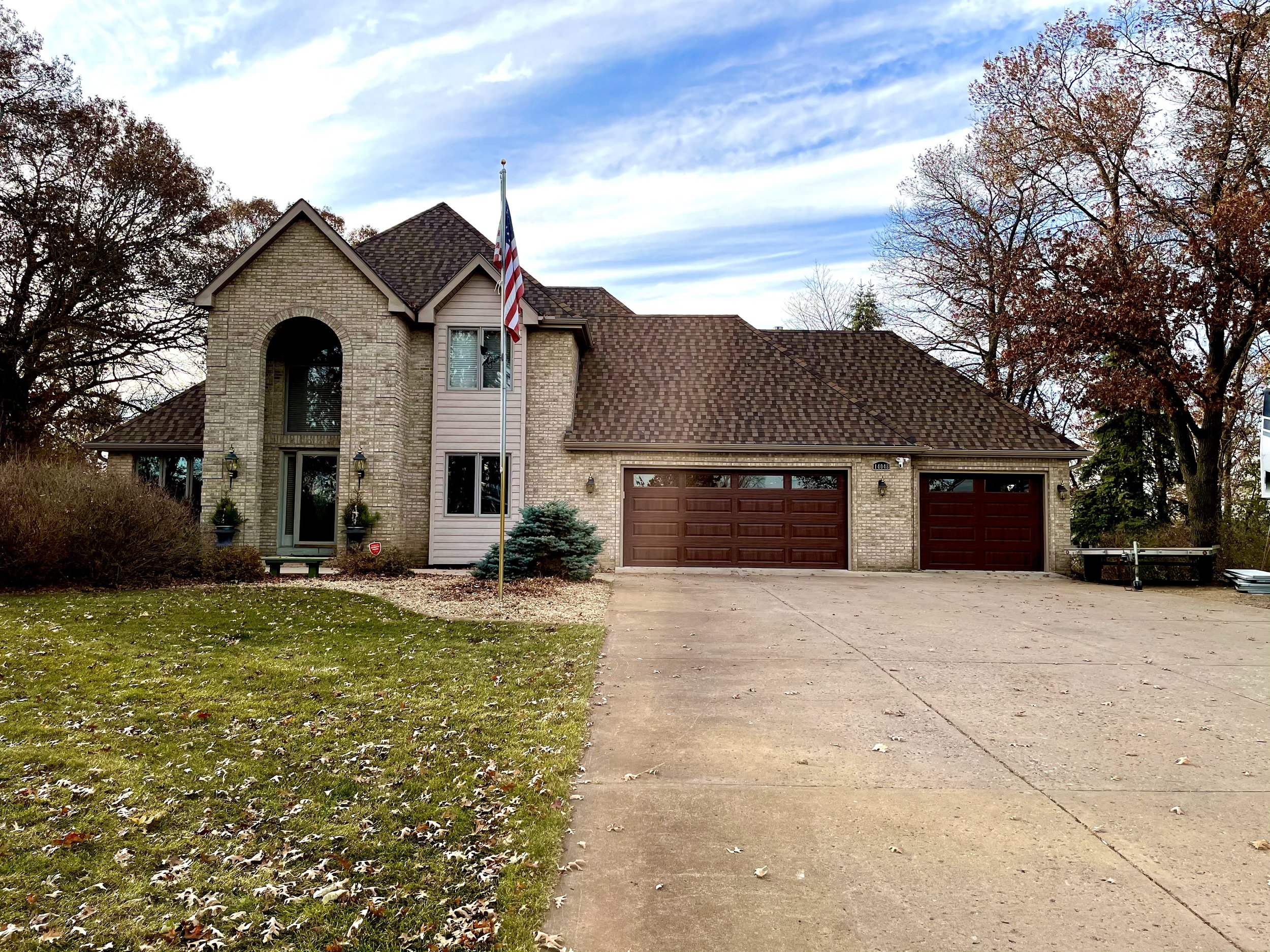 A large house with a brick facade, brown garage doors, and a two-story front entrance. The house has a manicured lawn, an American flag on a flagpole, and surrounding trees with autumn leaves.