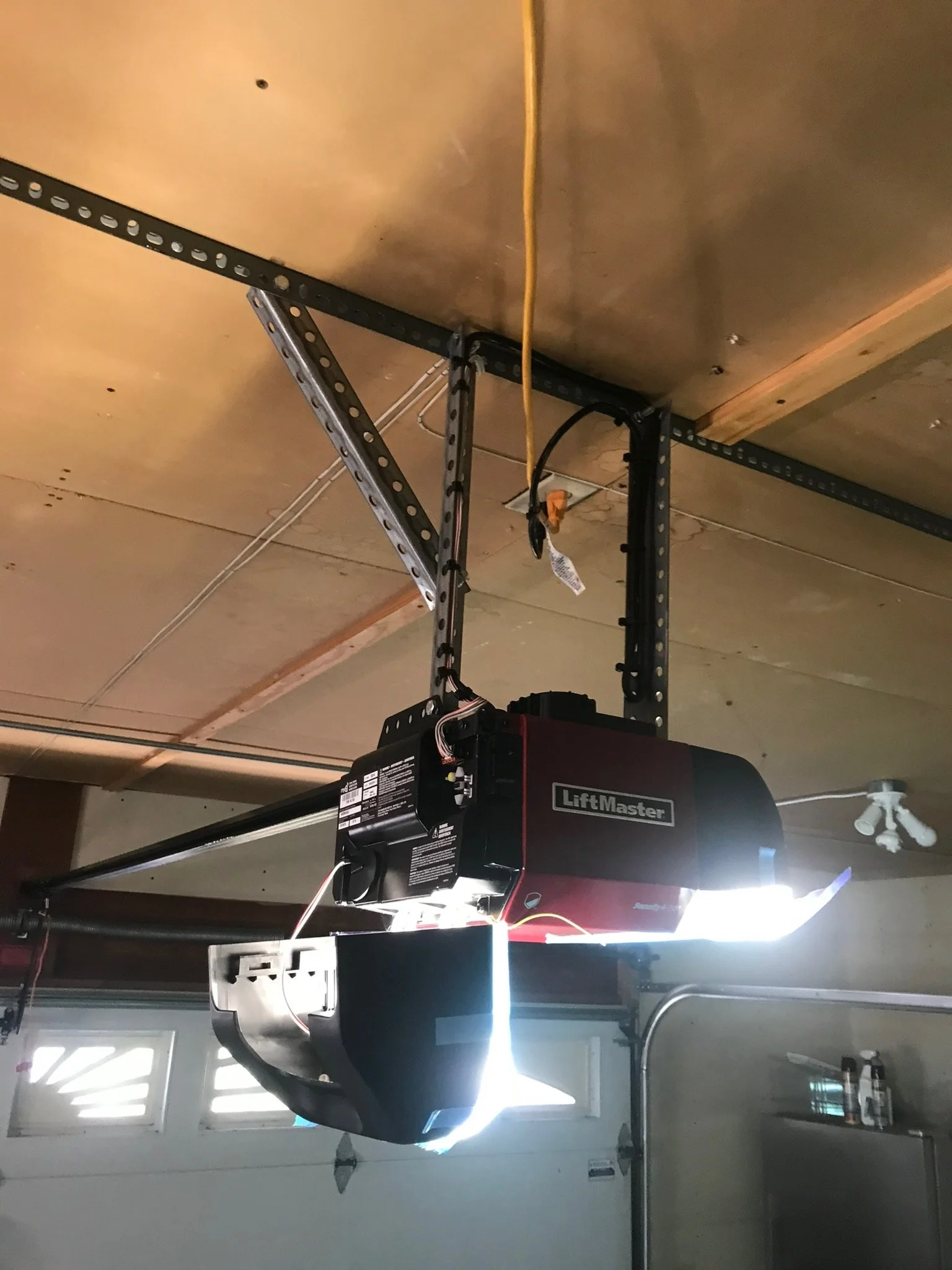 Garage door opener mounted on ceiling with exposed metal support tracks and electrical wiring, in a garage with windows and stored items.
