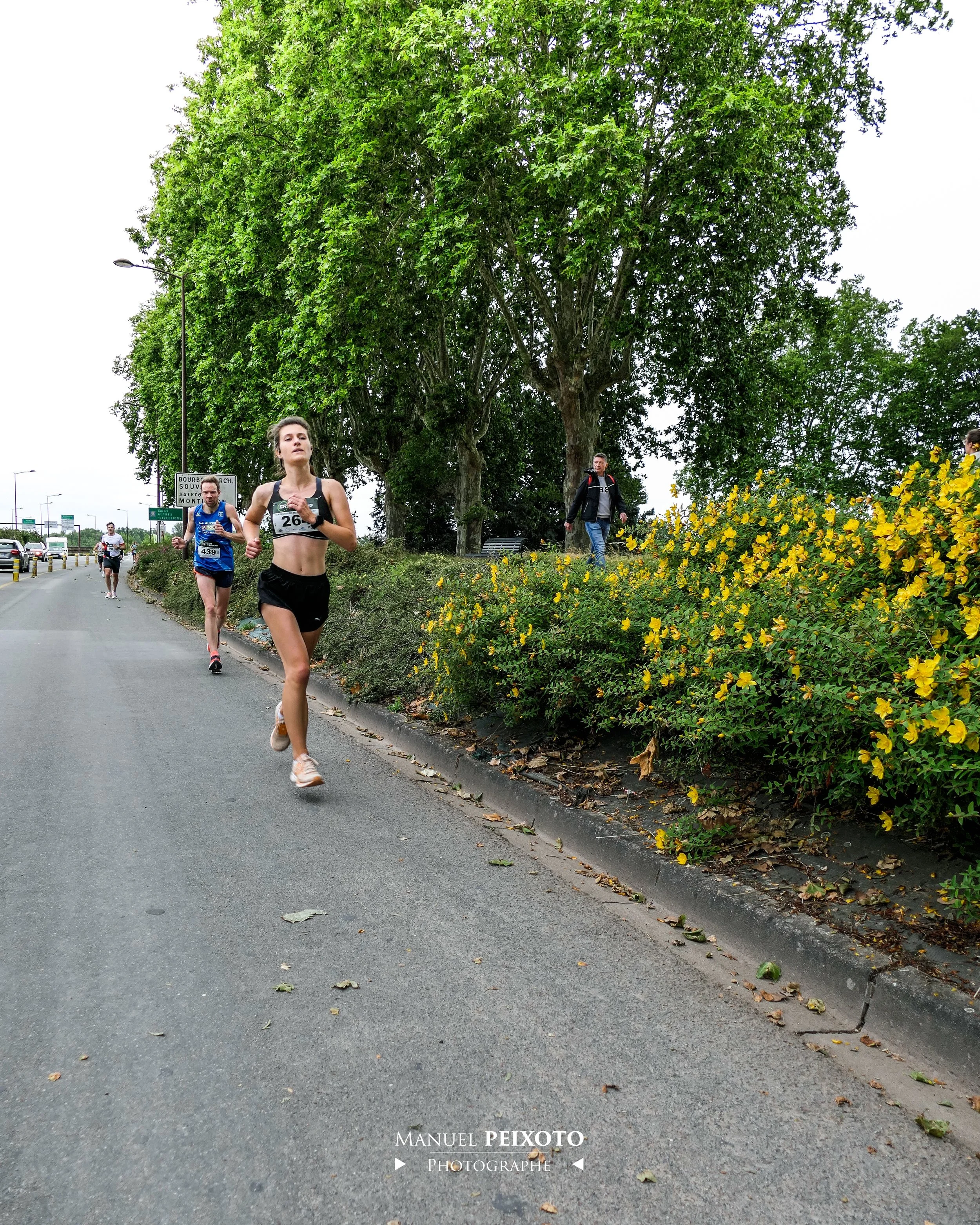 Des coureurs participant à une course de route, avec deux femmes en avant, l'une portant le numéro 26, sur un trottoir bordé d'arbres et de buissons jaunes. L'arrière-plan montre un ciel nuageux et une route avec des véhicules.