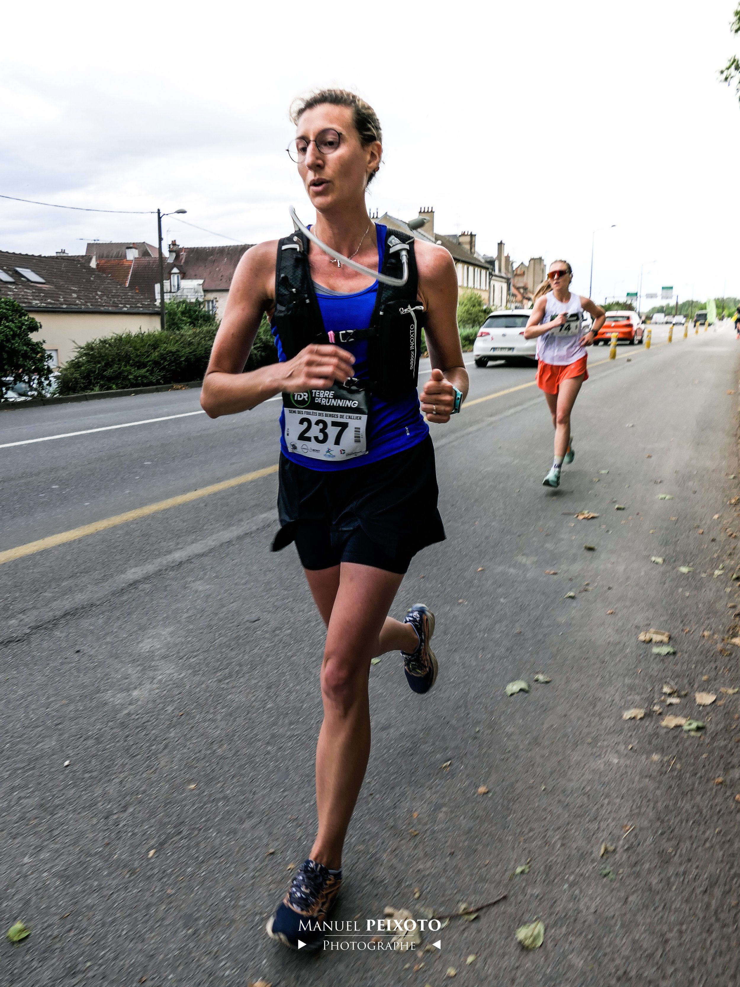 Deux femmes courent lors d'une course sur une route urbaine, l'une en avant portant un dossard 237 et une en arrière, toutes deux en tenue de course.