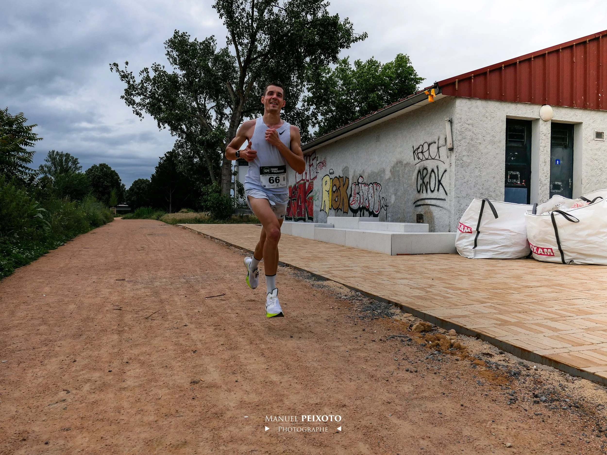 Un coureur en train de courir sur un chemin en extérieur sous un ciel nuageux, portant un dossard numéro 66, portant un t-shirt blanc, des baskets blanches, et des écouteurs.