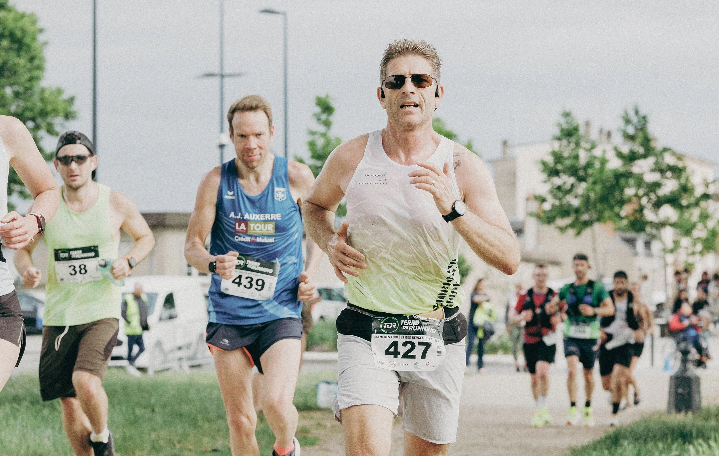 Groupe de coureurs participant à une course en plein air, portant des numéros d'identification et vêtus de vêtements de sport, avec un paysage urbain et des arbres en arrière-plan.