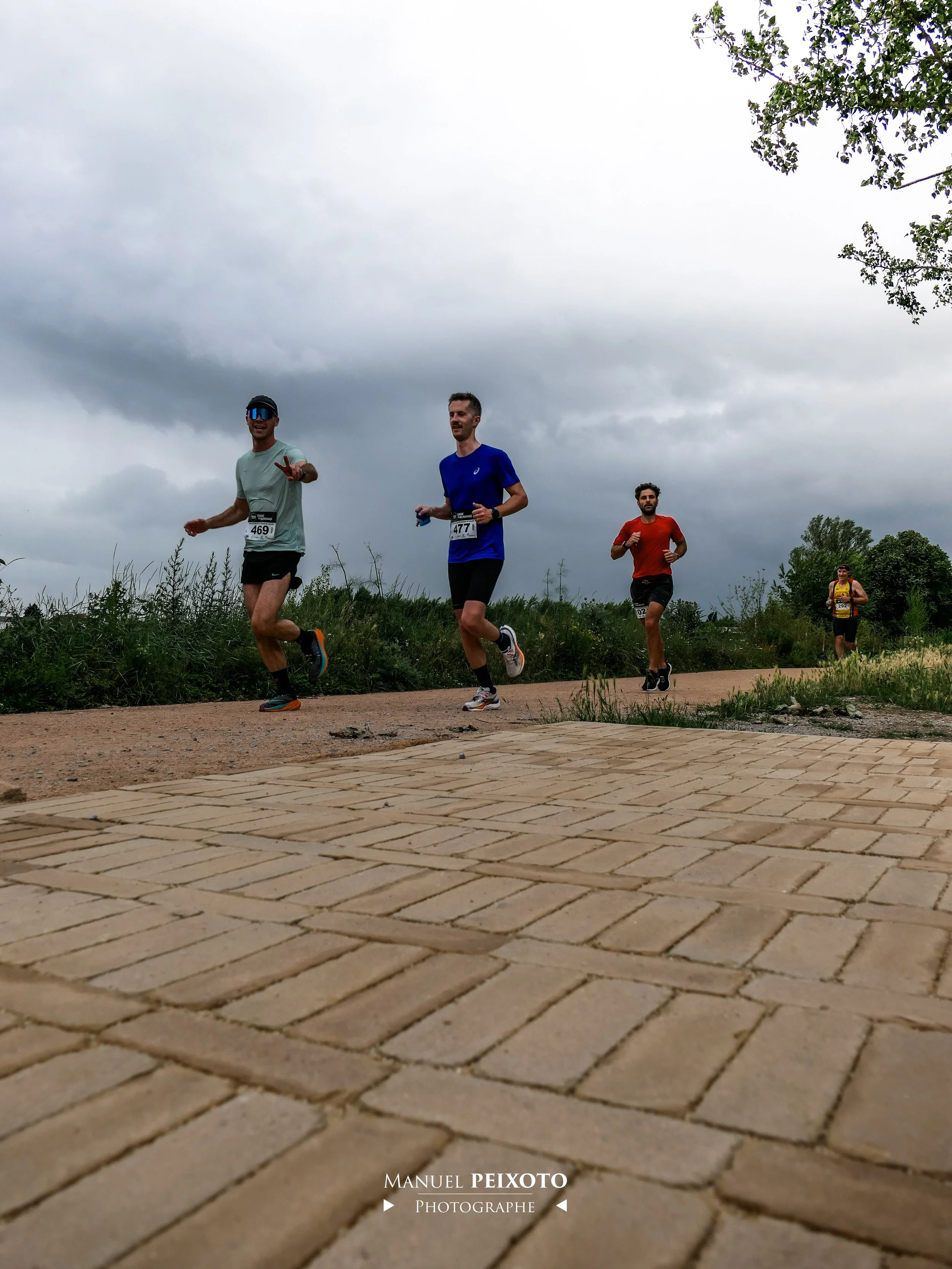 Quatre coureurs passent lors d'une course en extérieur sous un ciel nuageux, avec des panneaux en arrière-plan et un chemin pavé en premier plan.