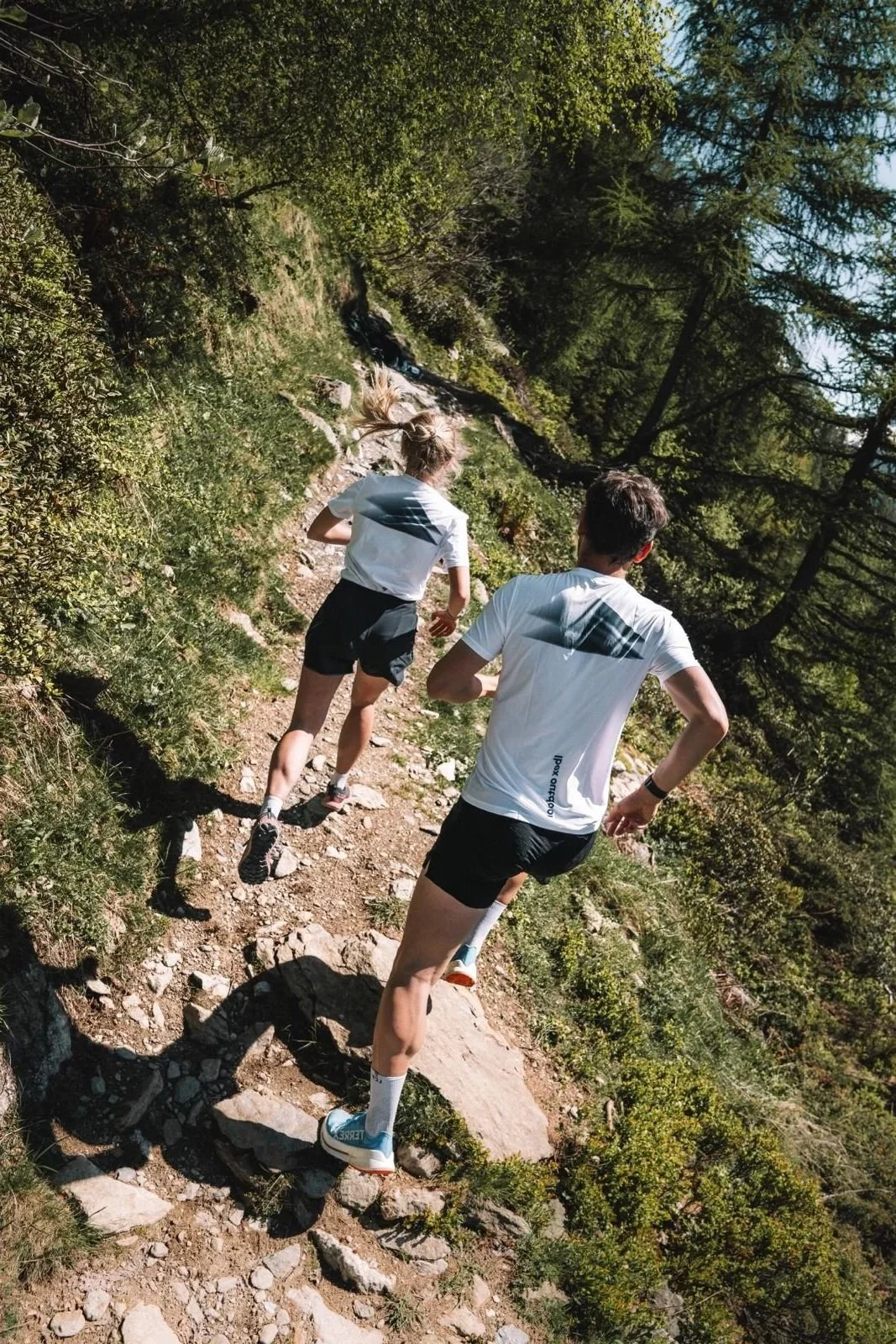 Deux personnes courent sur un sentier de montagne bordé d'arbres et de végétation, sous un ciel bleu clair.