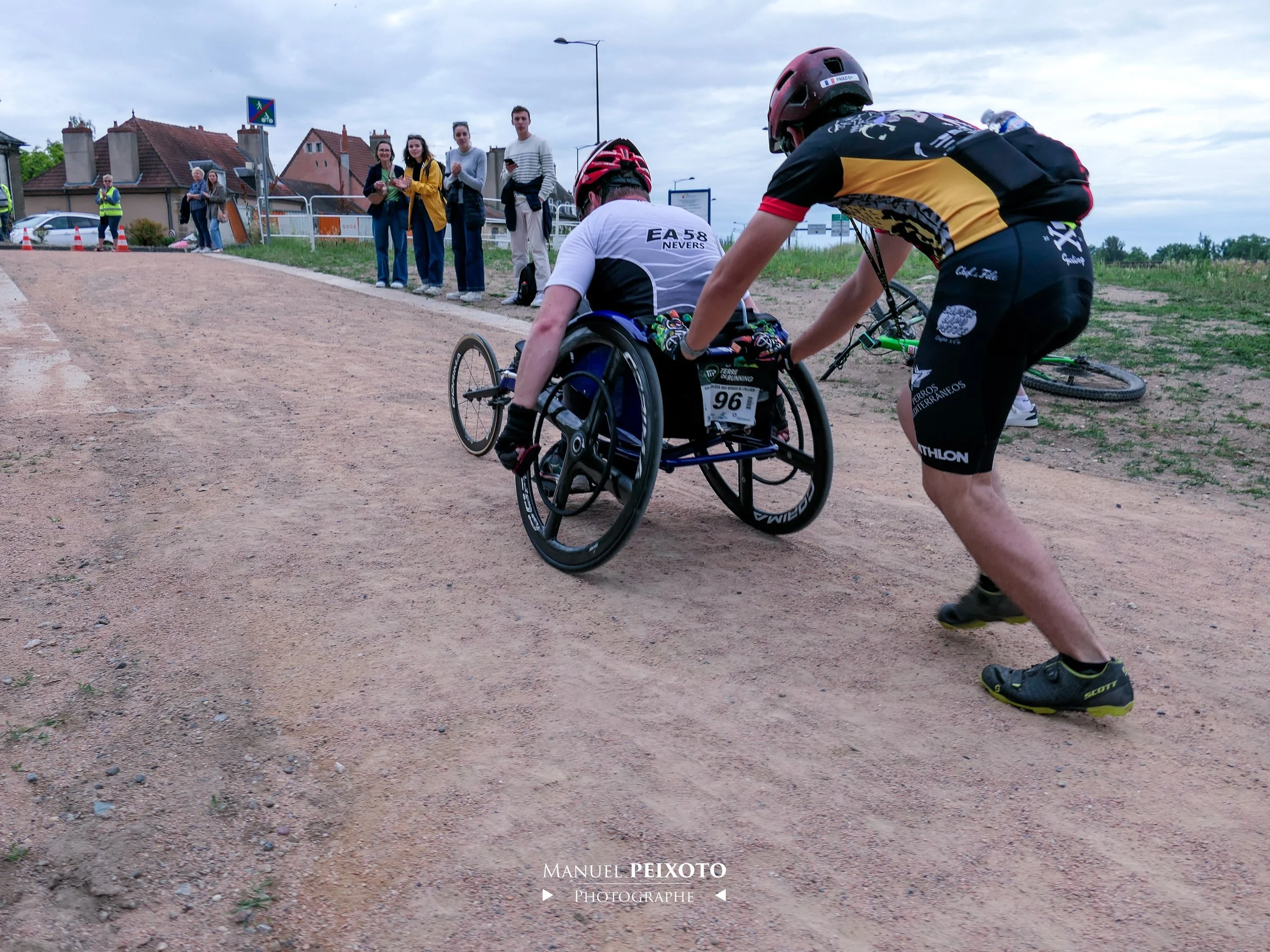 Un athlète en fauteuil roulant participant à une course, aidé par un supporteur, avec des spectateurs en arrière-plan