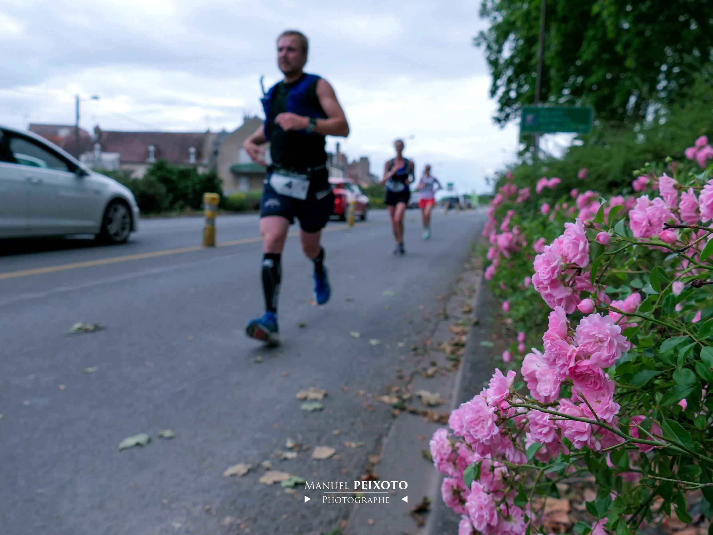 Trois coureurs participent à une course en plein air dans une ville. Le premier, proche de l'appareil, porte un maillot noir et des shorts, et court sur une piste pavée, avec des fleurs roses à sa droite. Deux autres coureurs sont visibles en arrière-plan, sous un ciel nuageux.