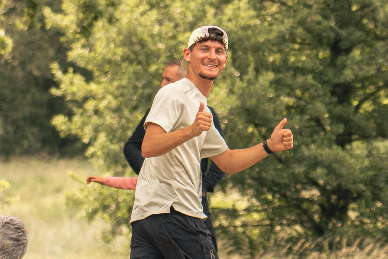 Jeune homme souriant donnant un pouce en l'air lors d'une activité de course en plein air, avec un fond de végétation verte.