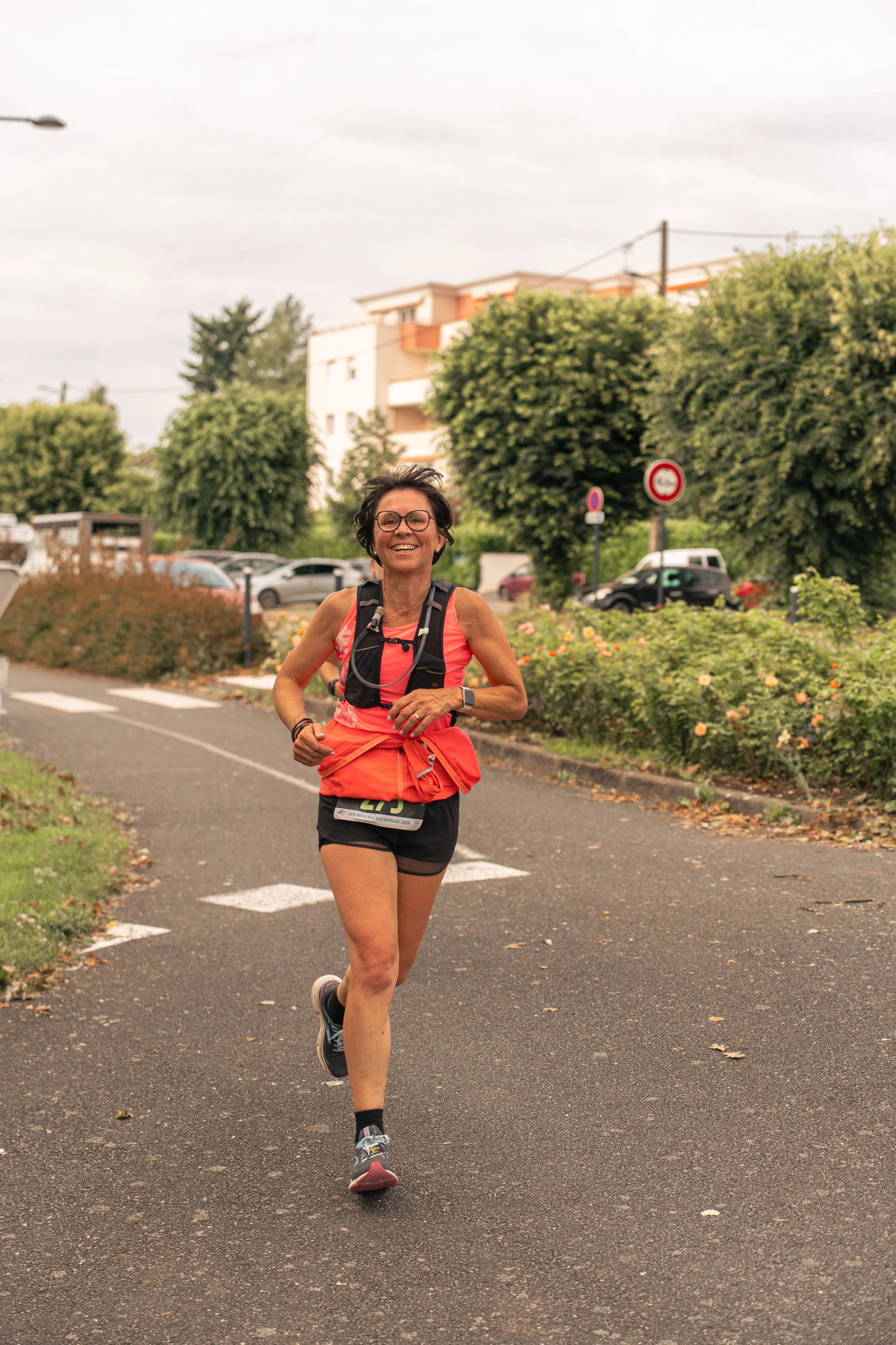 Une femme courent lors d'une course en plein air, portant une tenue de course orange et des lunettes, souriant.