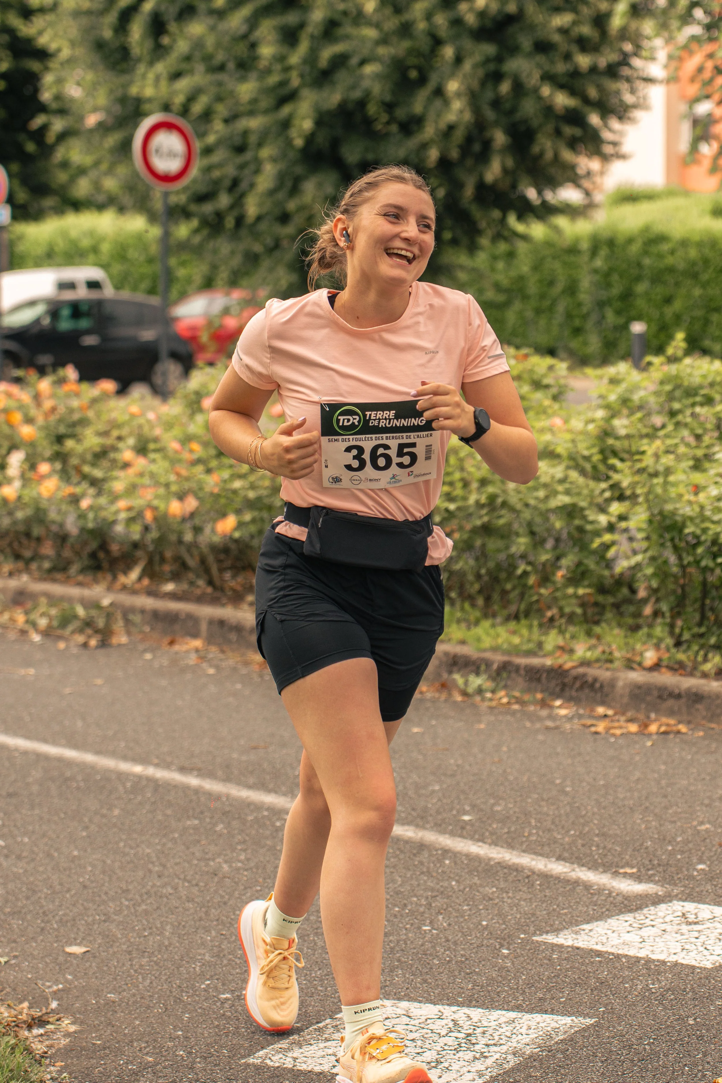Une femme souriante court lors d'une course, portant un dossard numéro 365, une tenue sportive rose et noire, et une montre bracelets, en arrière-plan des arbres et voitures.