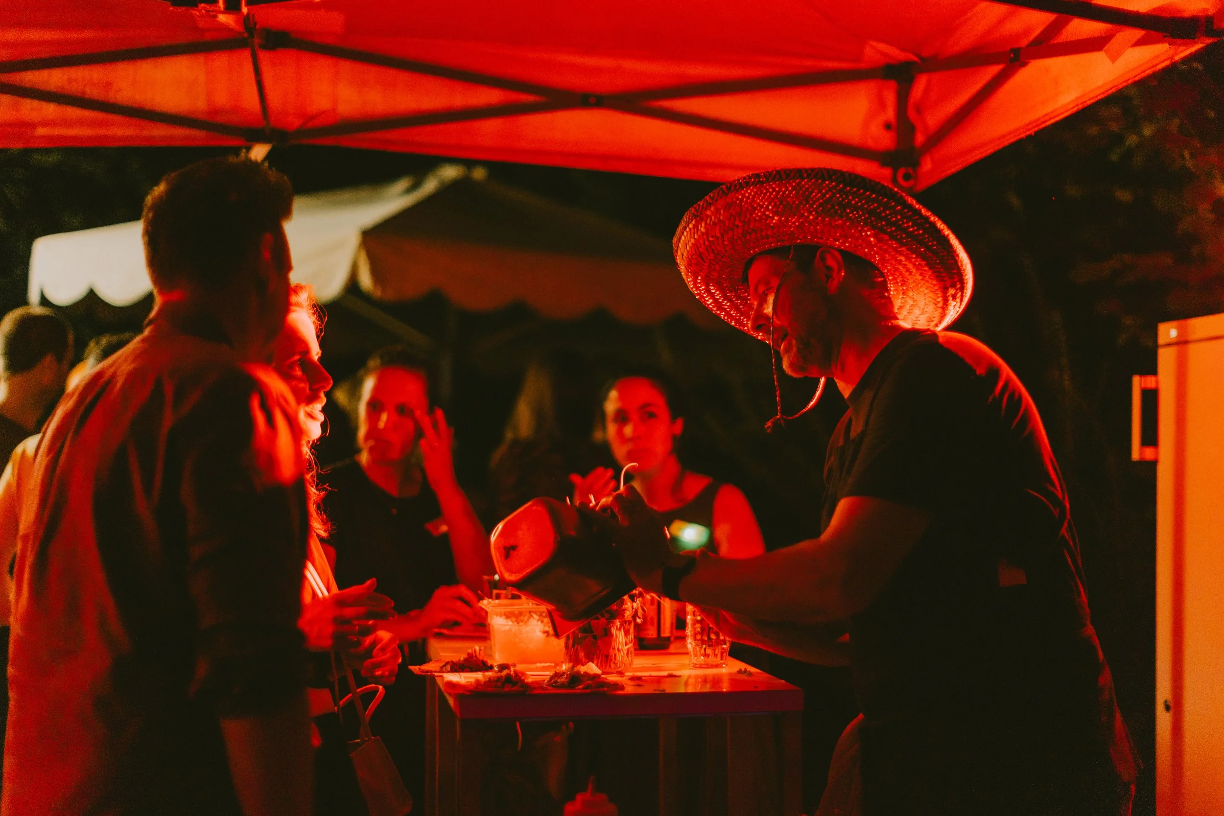 Menschen, die um einen Stand bei Nacht an einer Party oder einem Fest trinken, mit einem Mann im Sombrero, der Getränke serviert. Die Szene wird von rotem Licht beleuchtet.