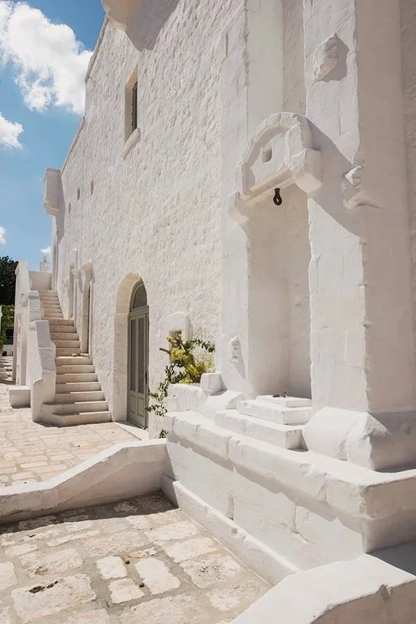 White stone building with stairs, a doorway, and a fountain or niche, located in a sunny outdoor area with cloudy sky.