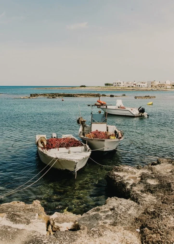 Two small boats docked near rocky shoreline with fishing nets, calm blue water, and a distant shoreline with white buildings.