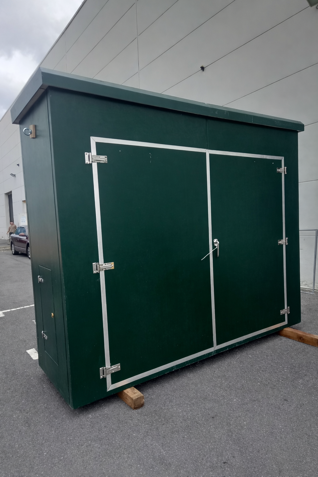 A large, dark green, metal storage shed with double doors secured by a lock and latch, placed outdoors on wooden supports, against a modern building with gray panels.